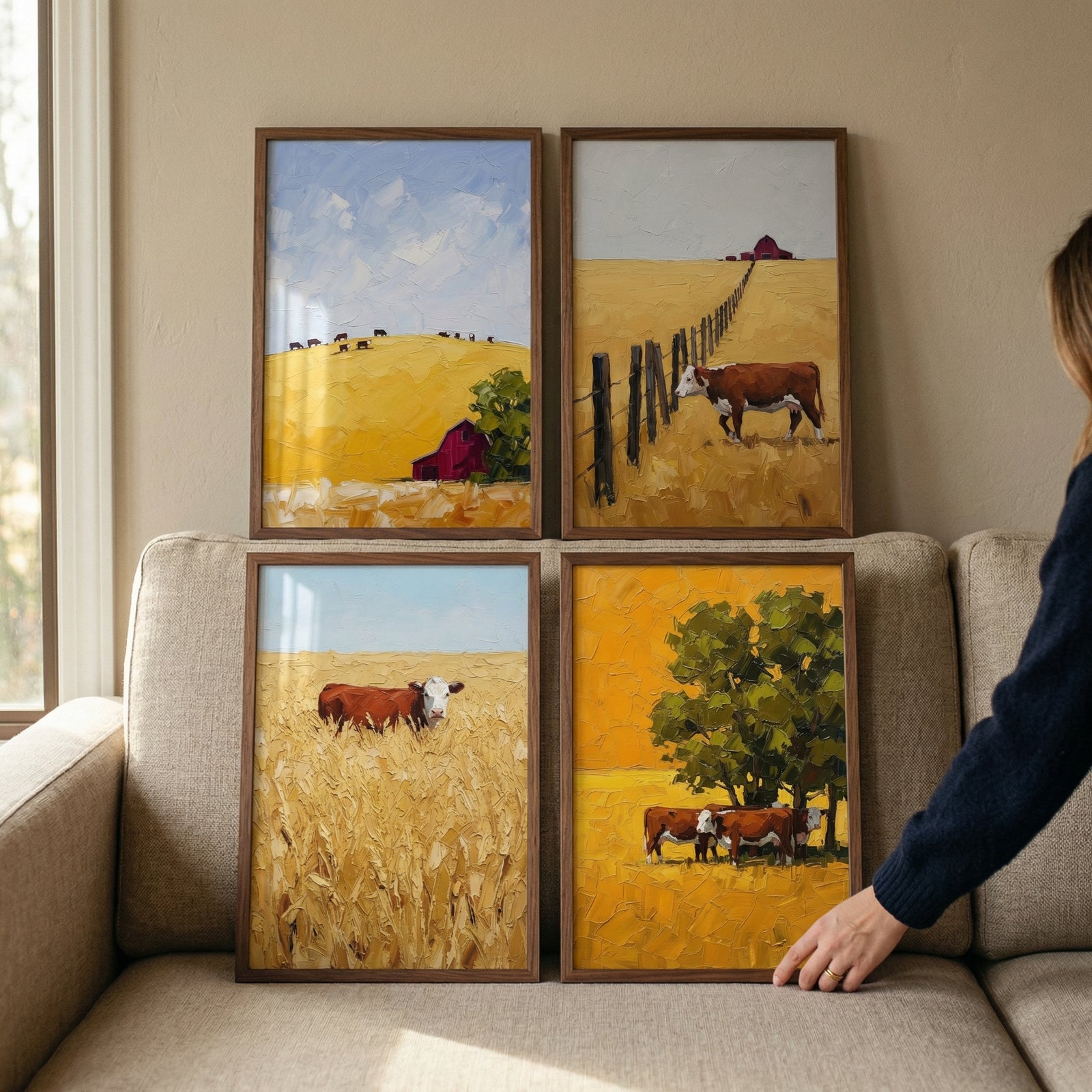 Four framed works are arranged on a light-colored couch. Each piece features a textured, impressionistic style capturing rural farm scenes in warm, golden tones. The top-left shows a golden hill with a red barn. The top-right depicts a cow by a fence in a yellow field. The bottom-left captures a cow standing in tall grain, and the bottom-right shows cattle shaded by a large green tree. 
