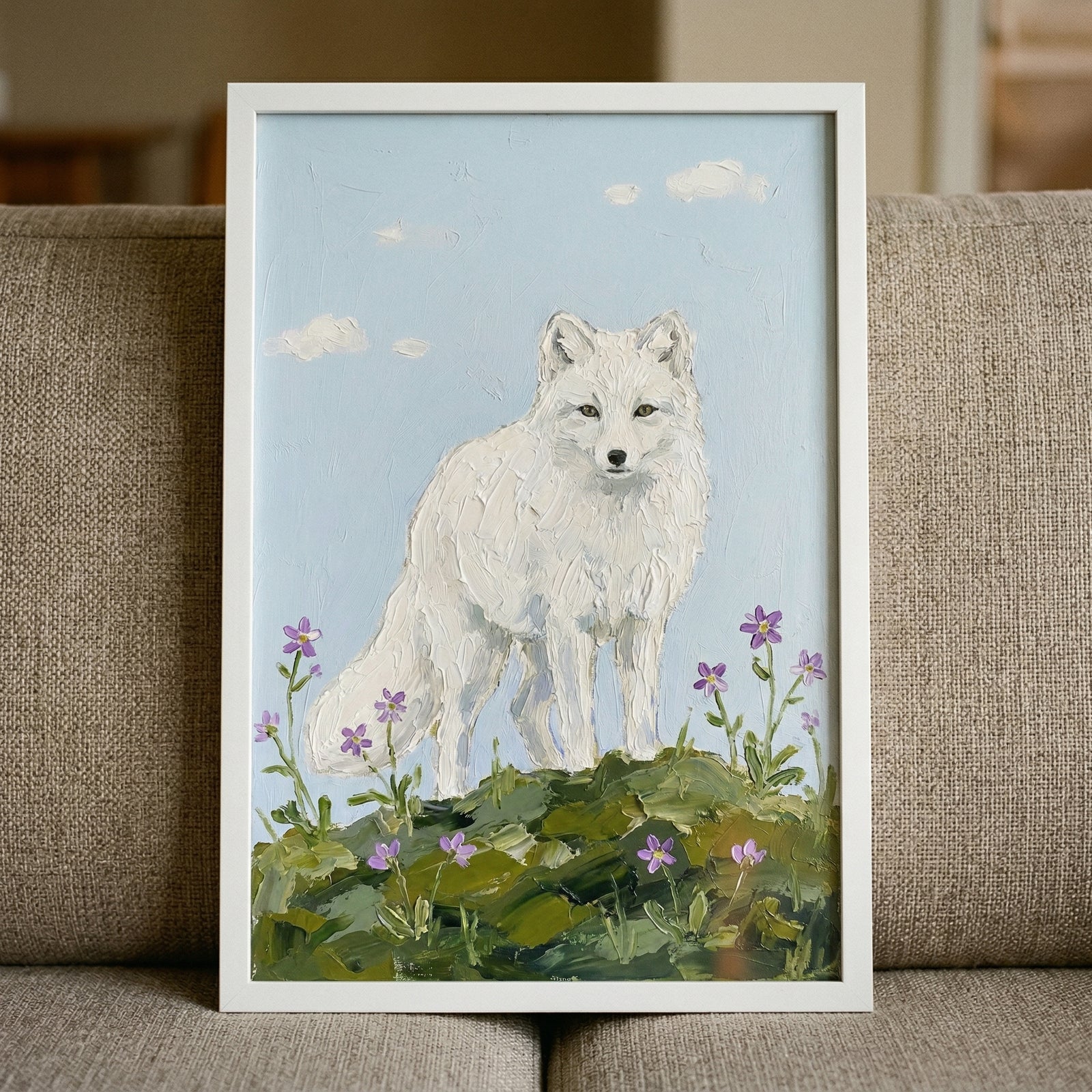 A textured artwork featuring a white arctic fox standing atop a small, grassy hill. The piece uses thick, visible brushstrokes to create a sense of depth and tactile surface, particularly in the fox's dense fur and the varied greens of the mound. Small, delicate purple wildflowers grow around the base of the animal. Above, the sky is a pale, calm blue with a few wispy white clouds. The entire creation is enclosed in a simple, thin white frame and is shown resting against the back of a grey fabric sofa.