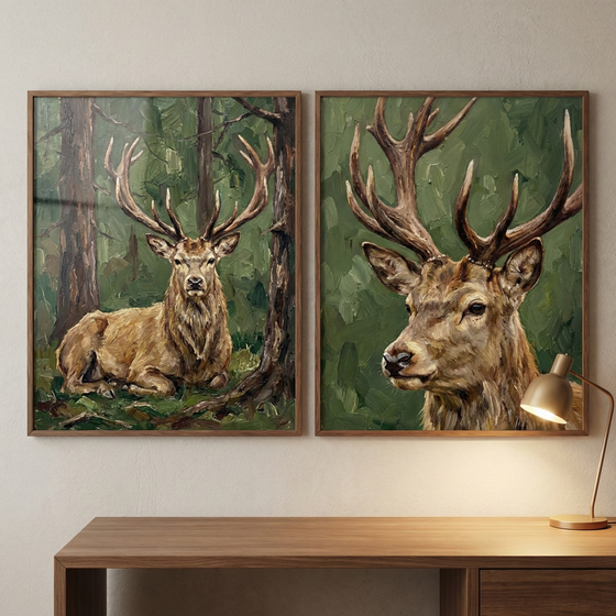 Two framed artworks hang side-by-side above a wooden desk. The left piece depicts a stag with large antlers resting in a forest of deep green and brown. The right piece is a close-up portrait of a stag's head, showcasing its impressive antlers against a textured green backdrop. Both images feature a bold, textured style. A small brass-colored desk lamp on the right casts a warm light. The matching dark wood frames complement the desk below.