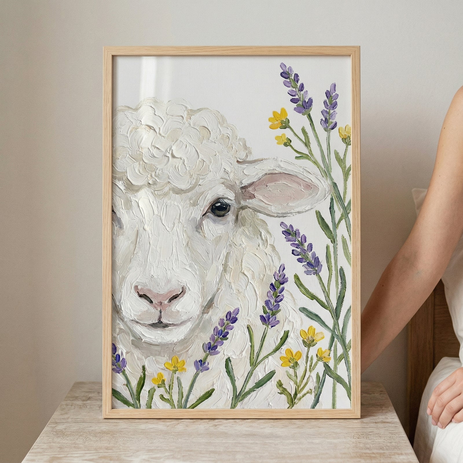 Close up painting of a white sheep with thick textured wool looking forward while surrounded by purple lavender stems and small yellow flowers. The artwork is displayed in a light wood frame on a table with a person seated beside it, highlighting its soft handcrafted detail.