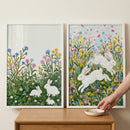 Two framed paintings of white rabbits sit on a wooden surface against a neutral wall, styled with a small dish and a person’s hand placing it nearby. The left artwork shows three rabbits nestled in green clover with pink, blue, and yellow flowers, while the right shows two rabbits leaping through a vibrant meadow filled with colorful blooms and scattered petals.