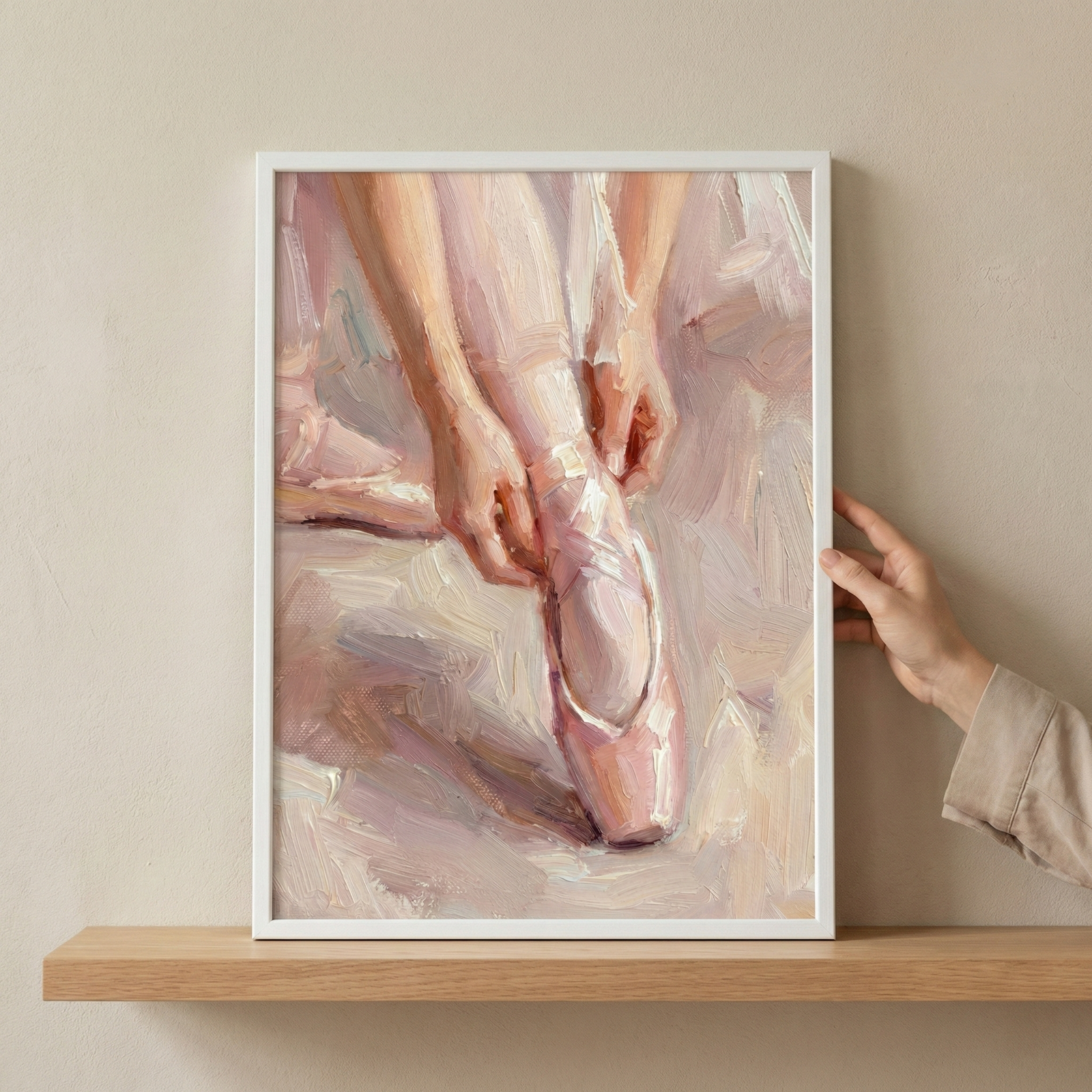 An impressionistic artwork depicts a ballet dancer's feet and lower legs. The dancer wears soft pink pointe shoes with delicate ribbons, and one hand is shown adjusting a shoe. The artwork features textured, visible strokes in a palette of cream, pale pink, and white. This piece is displayed in a white frame on a light-colored wooden shelf, with a person's hand visible on the right side holding the edge of the frame against a neutral-toned wall.