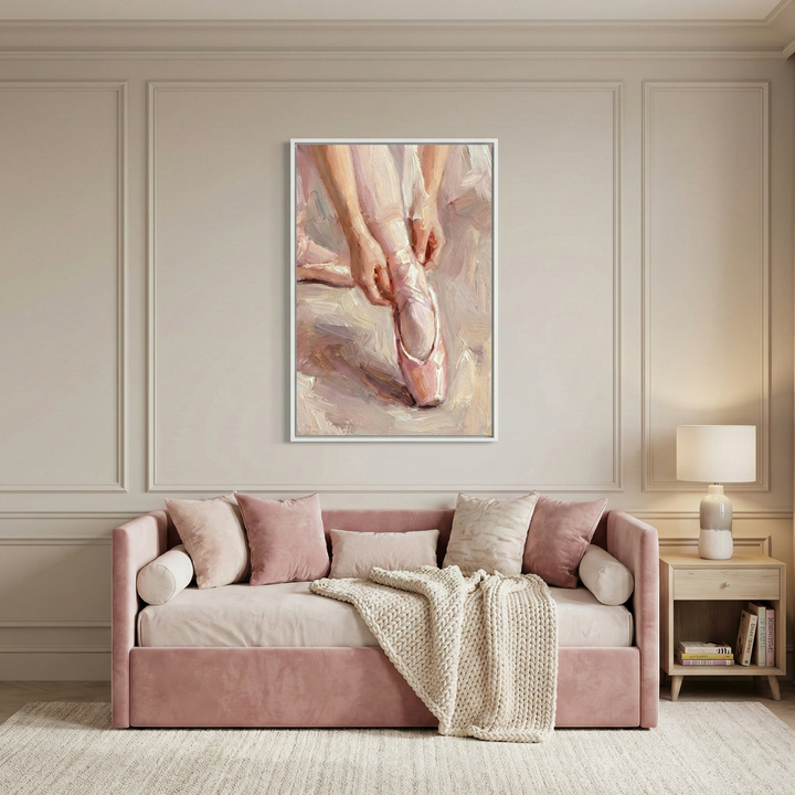 A cozy living room features a plush pink sofa adorned with soft cushions and a knitted throw. Above the sofa hangs a painting depicting a close-up of hands adjusting a ballet shoe. A wooden side table with a lamp and books completes the serene setting.