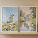 Two framed impressionist paintings leaning on a wooden shelf show a young blonde girl with a white horse in soft countryside scenes. One shows the girl riding the horse across a breezy wildflower hill while the other shows her leading the horse through a shallow reflective stream at sunset.