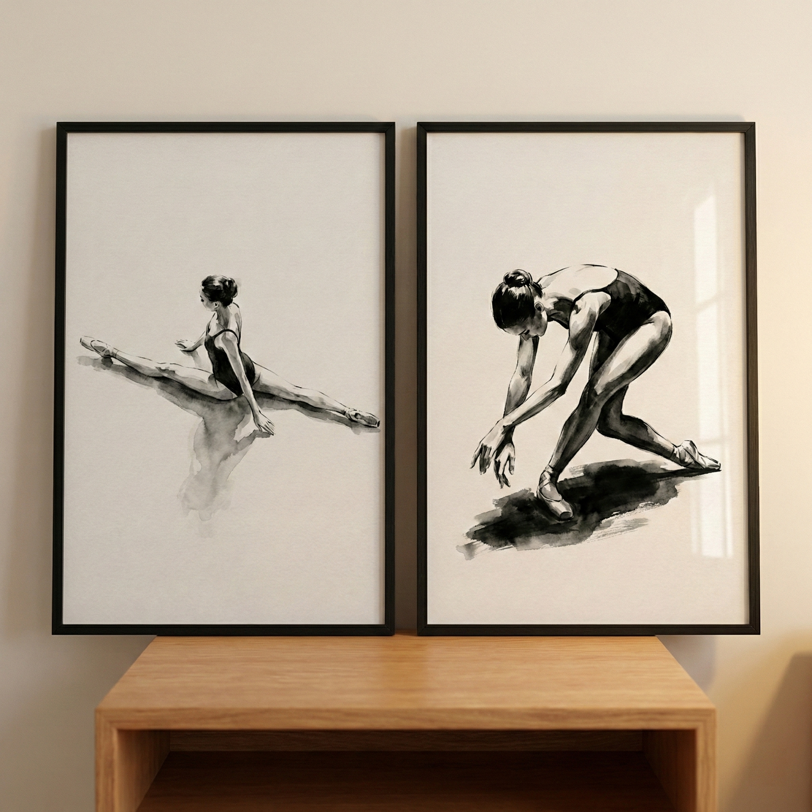 Two framed black and white drawings of a ballet dancer sit side-by-side on a wooden shelf against a plain wall. The piece on the left shows a dancer in a full split, her back to the viewer, with soft shadows underneath her. The piece on the right depicts the dancer in a dynamic pose, leaning forward with arms reaching down and one leg trailing behind, creating a sense of graceful movement. Both works use expressive brushwork and high contrast, set within simple black frames.