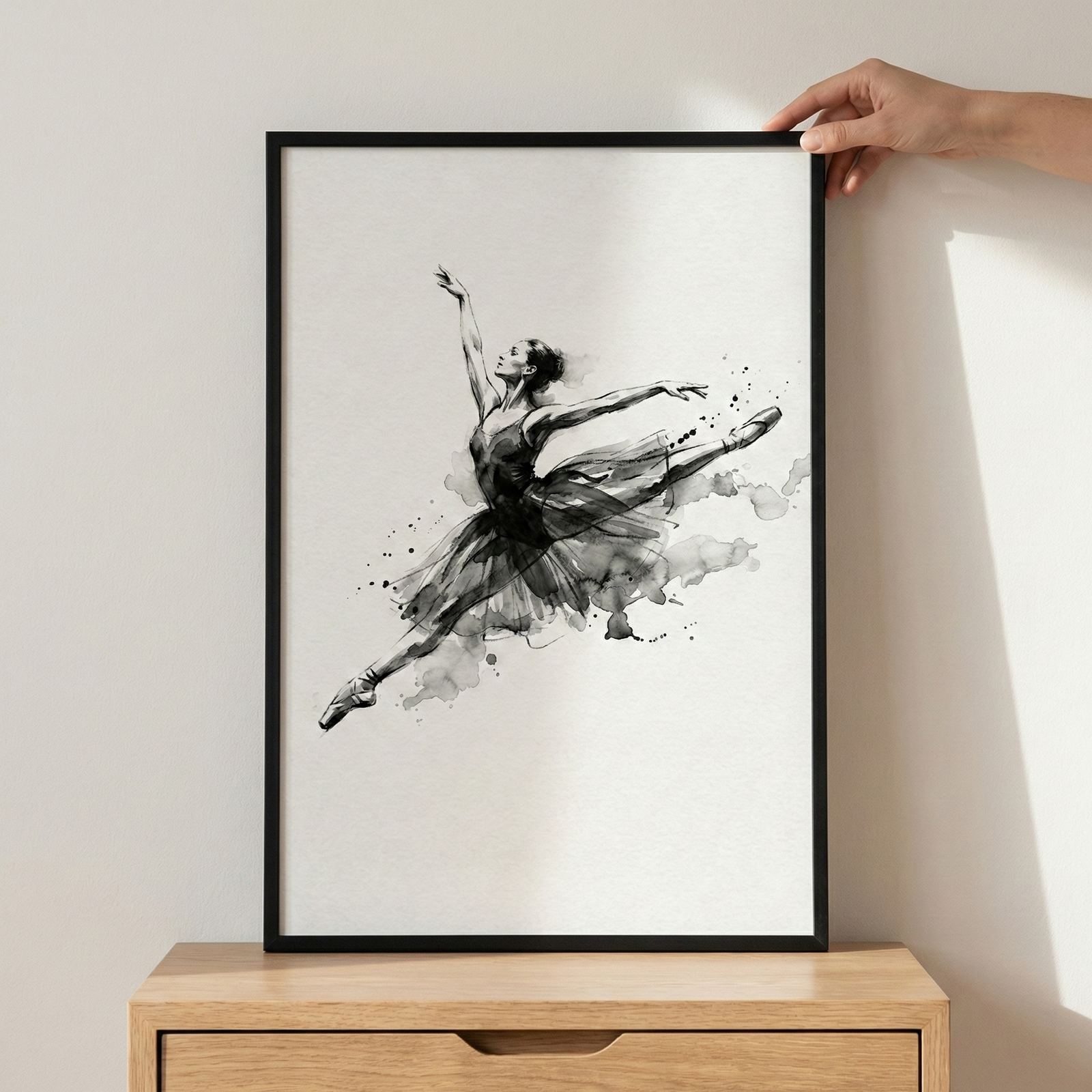 A monochrome ink-style artwork depicts a ballerina in mid-leap, her body extended in a graceful pose. The figure is rendered with expressive black strokes and soft grey washes, creating a dynamic sense of motion with artistic splatters and blurred edges around her dark tutu. This vertical artwork is housed in a simple black frame, held against a light-colored wall by a person's hand while resting atop a light wood dresser.