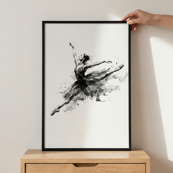 A monochrome ink-style artwork depicts a ballerina in mid-leap, her body extended in a graceful pose. The figure is rendered with expressive black strokes and soft grey washes, creating a dynamic sense of motion with artistic splatters and blurred edges around her dark tutu. This vertical artwork is housed in a simple black frame, held against a light-colored wall by a person's hand while resting atop a light wood dresser.