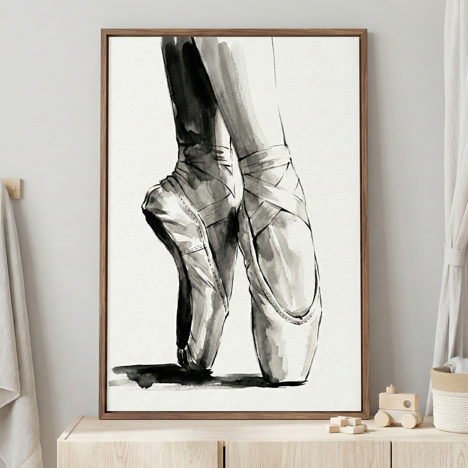 A framed monochromatic ink wash illustration depicts a dancer's feet en pointe. The artwork uses fluid black and gray brushstrokes to show the intricate ribbons and satin-like texture of ballet shoes. Set in a natural wood frame, the piece rests on a light-colored dresser in a bright, minimalist room. To its right are small wooden toys and a woven basket, while a soft white towel and gauzy curtain complete the peaceful, modern setting.