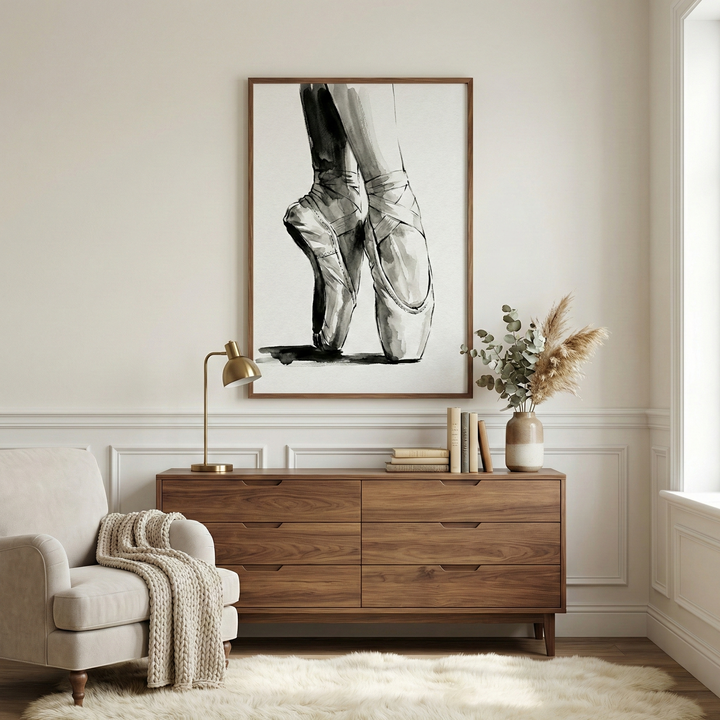 A serene room interior is shown. In the center, a large framed wall art piece depicts a pair of dancer's feet in pointe shoes, rendered in a black and white expressive sketch style. The wooden frame is a warm, medium brown. Below the art is a matching wooden six-drawer dresser. On the dresser to the left sits a gold desk lamp with a rounded shade. In the middle are a few stacked books, and to the right is a beige and cream ceramic vase filled with dried eucalyptus and pampas grass.