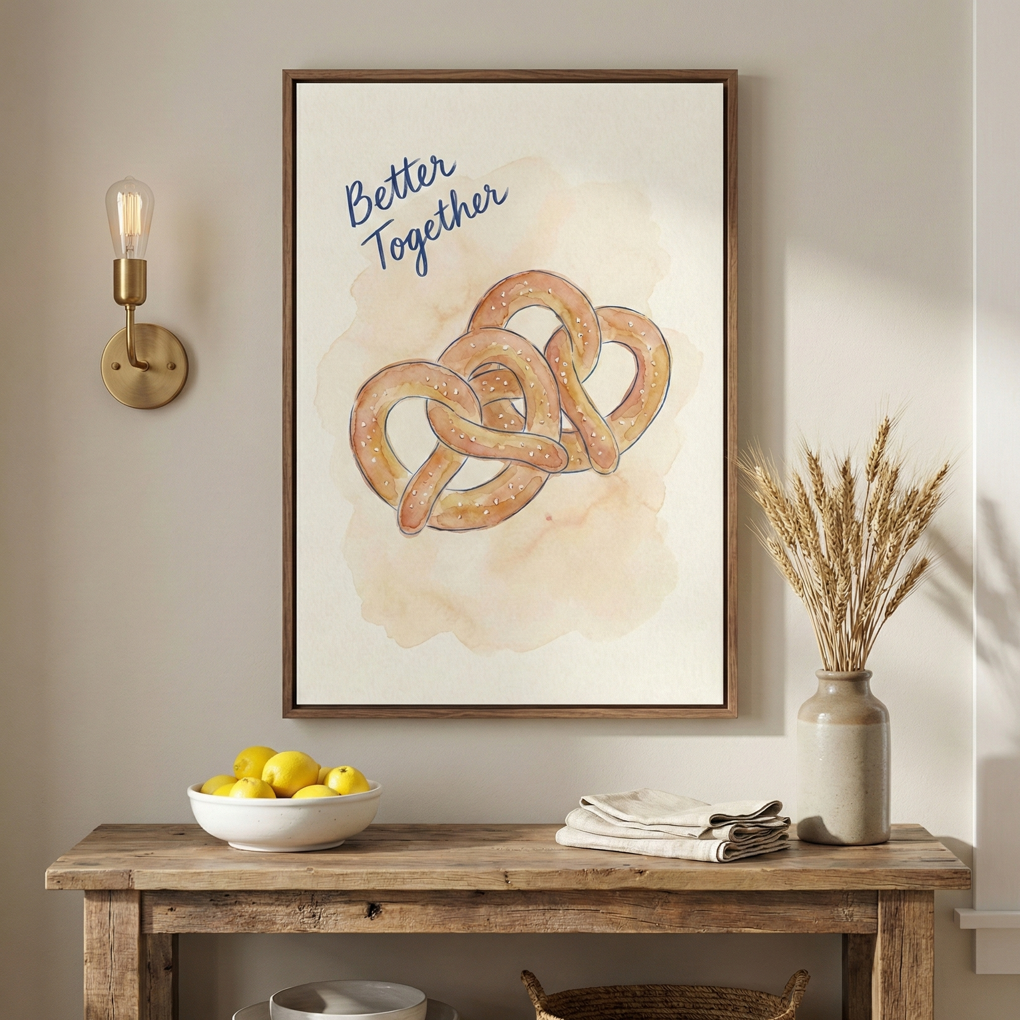 A framed watercolor-style illustration of two intertwined pretzels with the phrase "Better Together" written in blue cursive above them. The artwork is mounted on a light beige wall above a rustic wooden console table. On the table, there is a white bowl filled with lemons, a stack of folded off-white napkins, and a beige ceramic vase containing dried wheat. A brass wall sconce with an exposed bulb is visible on the wall to the left of the frame.