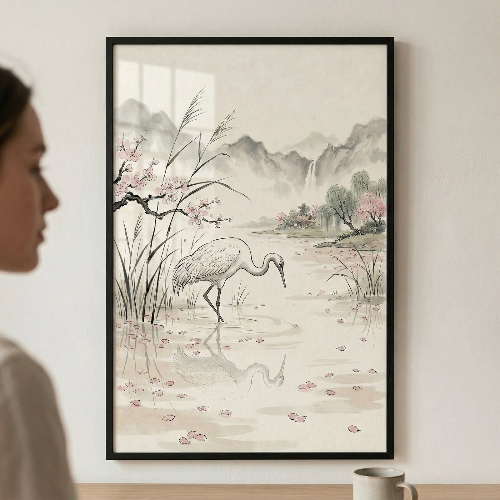 A vertical framed artwork hangs on a light-colored wall. The illustration, in an East Asian ink-and-wash style, features a white crane standing in shallow water, its neck curved as it looks down at its reflection. Pink petals are scattered across the water's surface. On the left, tall reeds and a blossoming branch with small pink flowers frame the bird. In the background, hazy mountains and a waterfall are rendered in soft grey and green tones under a pale sky. 