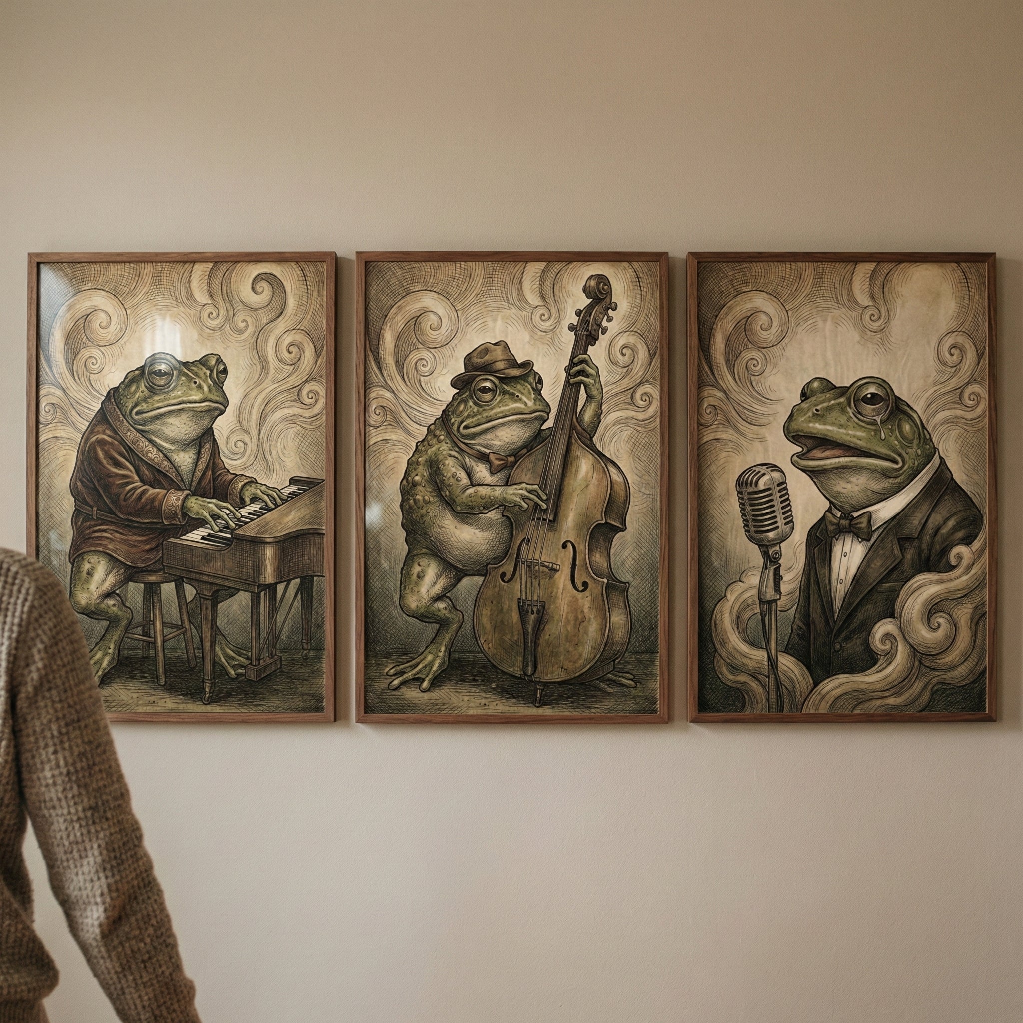 Three wood-framed illustrations of anthropomorphic frogs as jazz musicians hang on a neutral-colored wall. On the left, a frog wearing a smoking jacket and monocle plays a piano. In the center, a frog in a fedora and bow tie plays an upright bass. On the right, a frog in a tuxedo and bow tie sings into a vintage-style microphone. The artwork features a vintage, sepia-toned style with swirling smoke-like patterns in the background. A person's shoulder in a brown knit sweater is partially visible on the left.