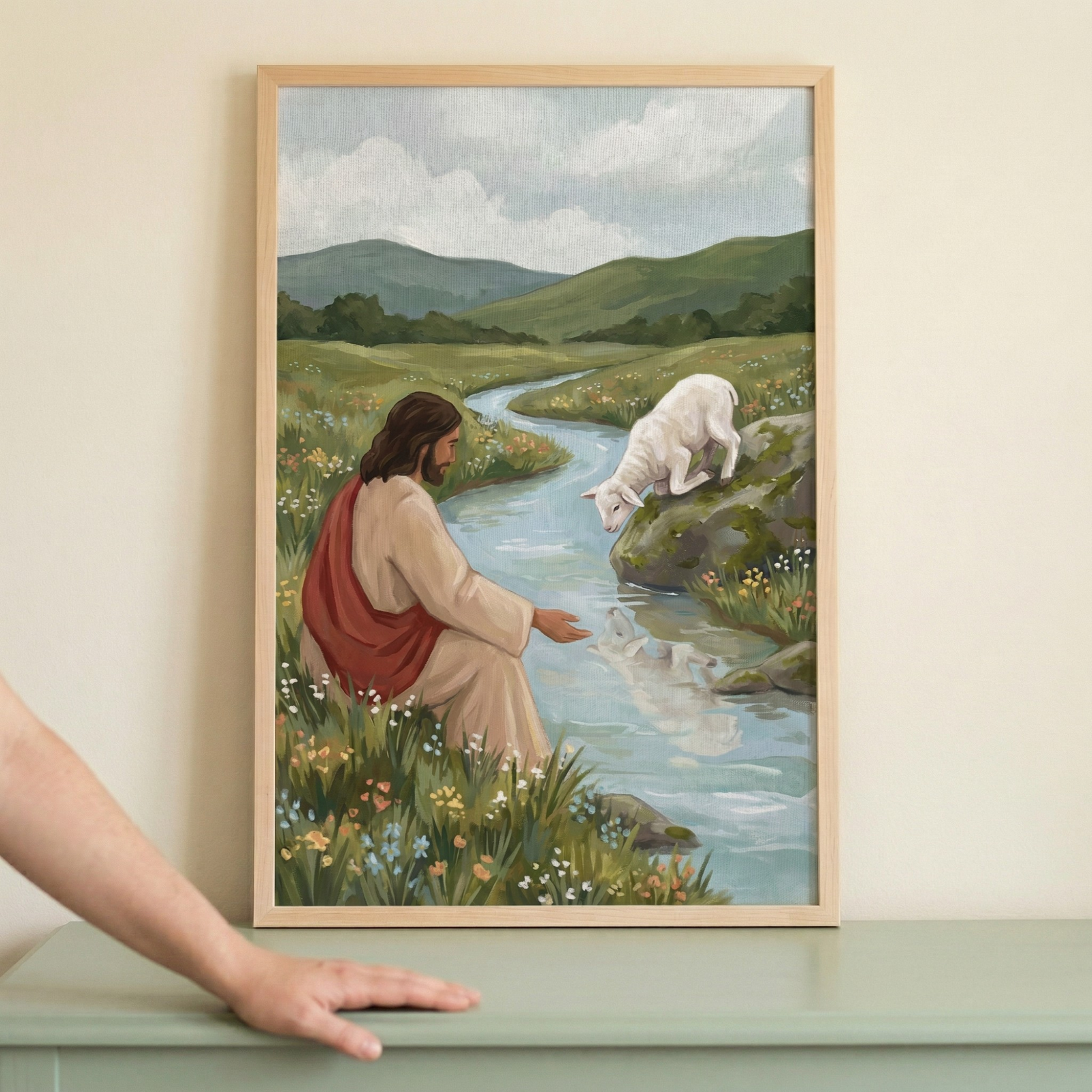 A framed illustration depicting Jesus Christ sitting peacefully on a grassy bank by a winding stream. On the opposite side of the water, a small white lamb bows its head to drink. The landscape features rolling green hills under a soft, cloudy sky. Jesus is shown with long brown hair, wearing a tan robe and a red sash. The scene is filled with wildflowers and soft light, capturing a serene pastoral moment. The artwork is presented in a light-colored wooden frame.