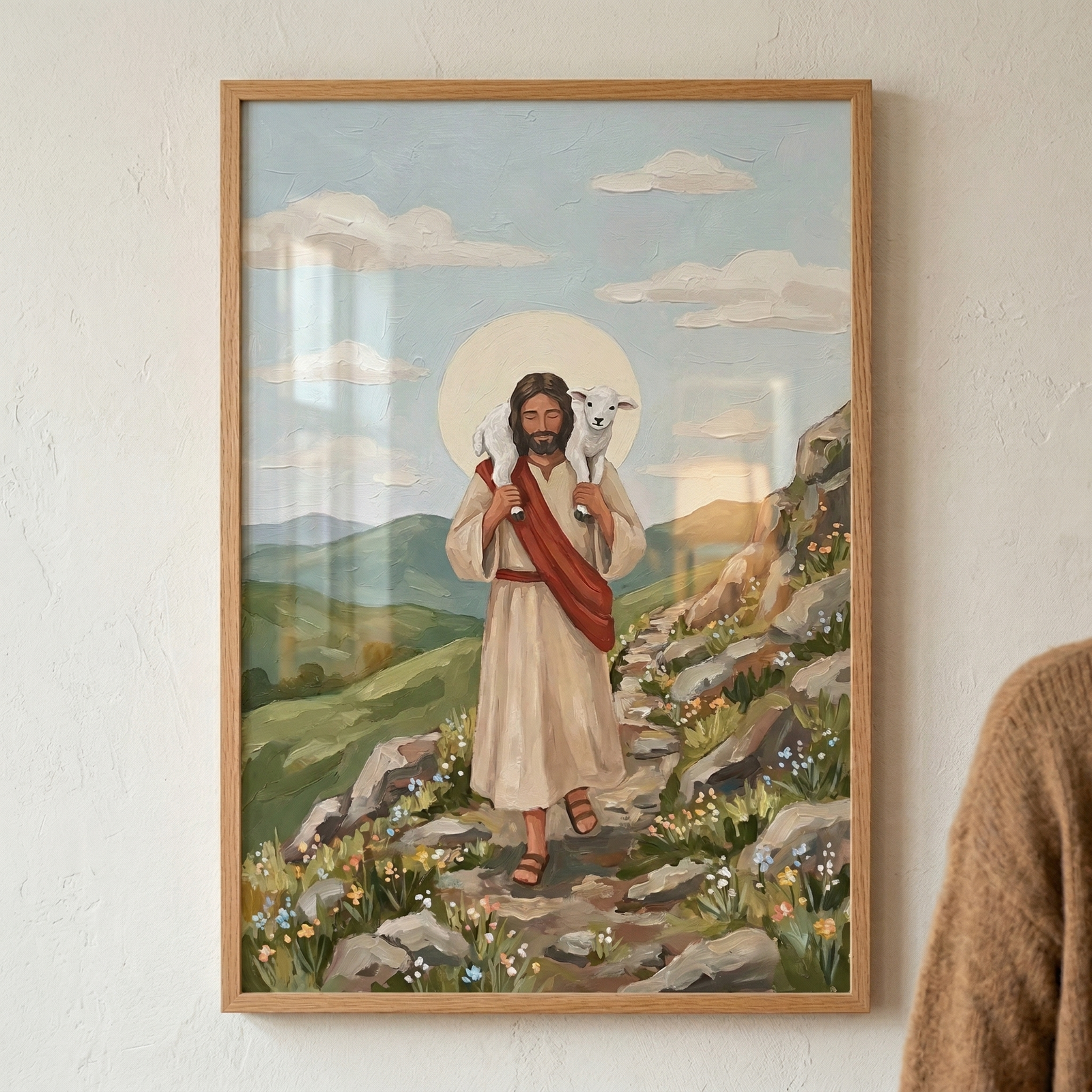 A vertical framed artwork depicts Jesus walking down a rocky mountain path, carrying a small white lamb across his shoulders. He wears a cream-colored robe with a red sash, and a soft yellow circular halo glows behind his head. The landscape features rolling green hills under a pale blue sky with light clouds. Tiny wildflowers in shades of blue and yellow grow along the trail. The illustration has a textured, expressive style with visible brushwork throughout the scene.