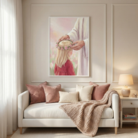 A framed artwork hangs on a light-colored wall with decorative molding above a white sofa. The artwork depicts Jesus Christ gently placing a crown of pink flowers on the head of a young girl with blonde hair who is wearing a red dress. The background of the illustration features a soft field of flowers under a pale pink sky. The sofa below is styled with several plush pillows in shades of dusty rose and cream, along with a thick, chunky knit beige throw blanket. 