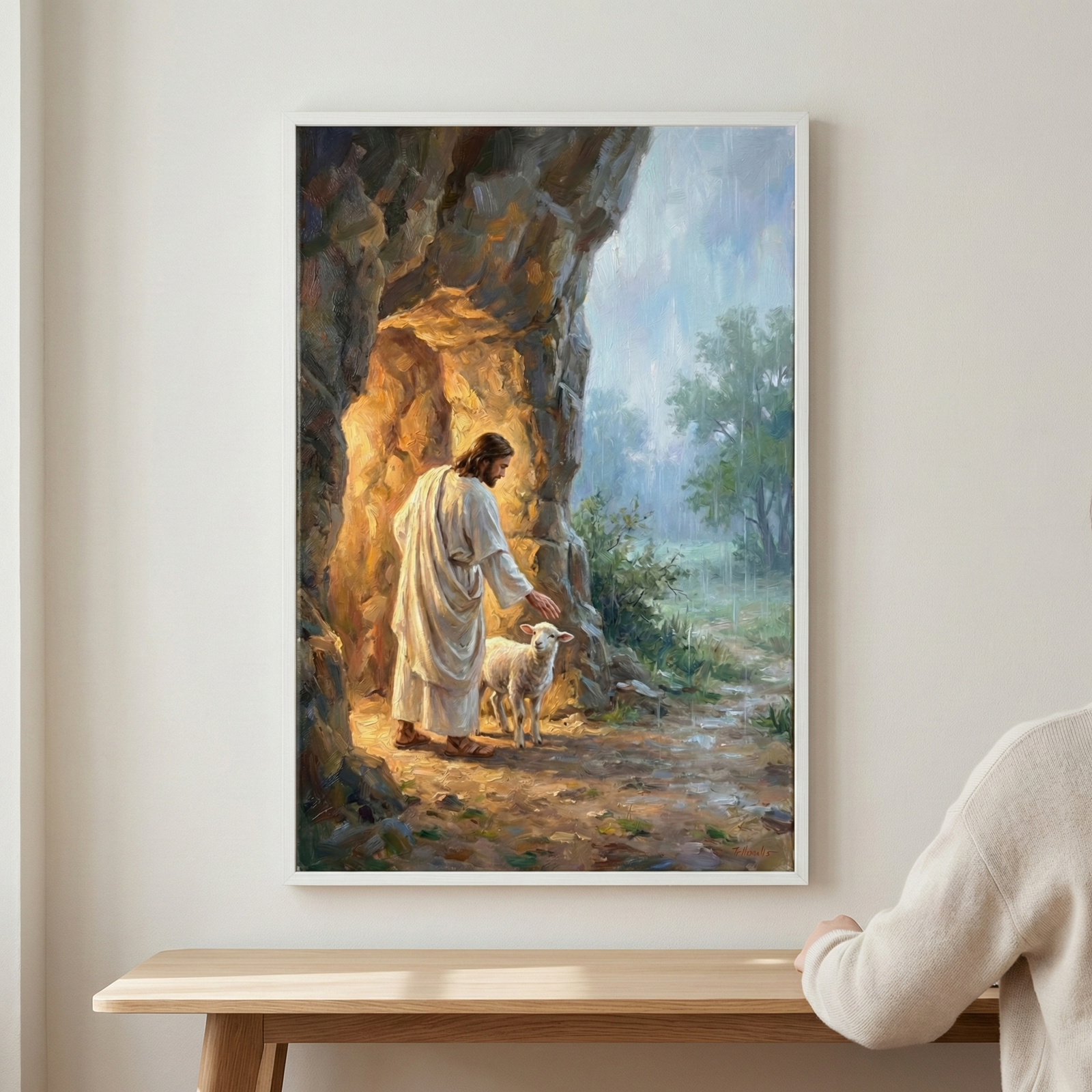 A vertical framed art piece hangs on a light-colored wall above a wooden table. The artwork depicts Jesus Christ dressed in a white robe, standing at the mouth of a sunlit cave. He gently rests his hand on a small white lamb. Warm, golden light spills from the cave's interior, contrasting with the soft, misty rain falling on the green landscape and trees in the background. The art features visible brushwork and rich textures. A portion of a person's shoulder in a beige sweater is seen on the right.