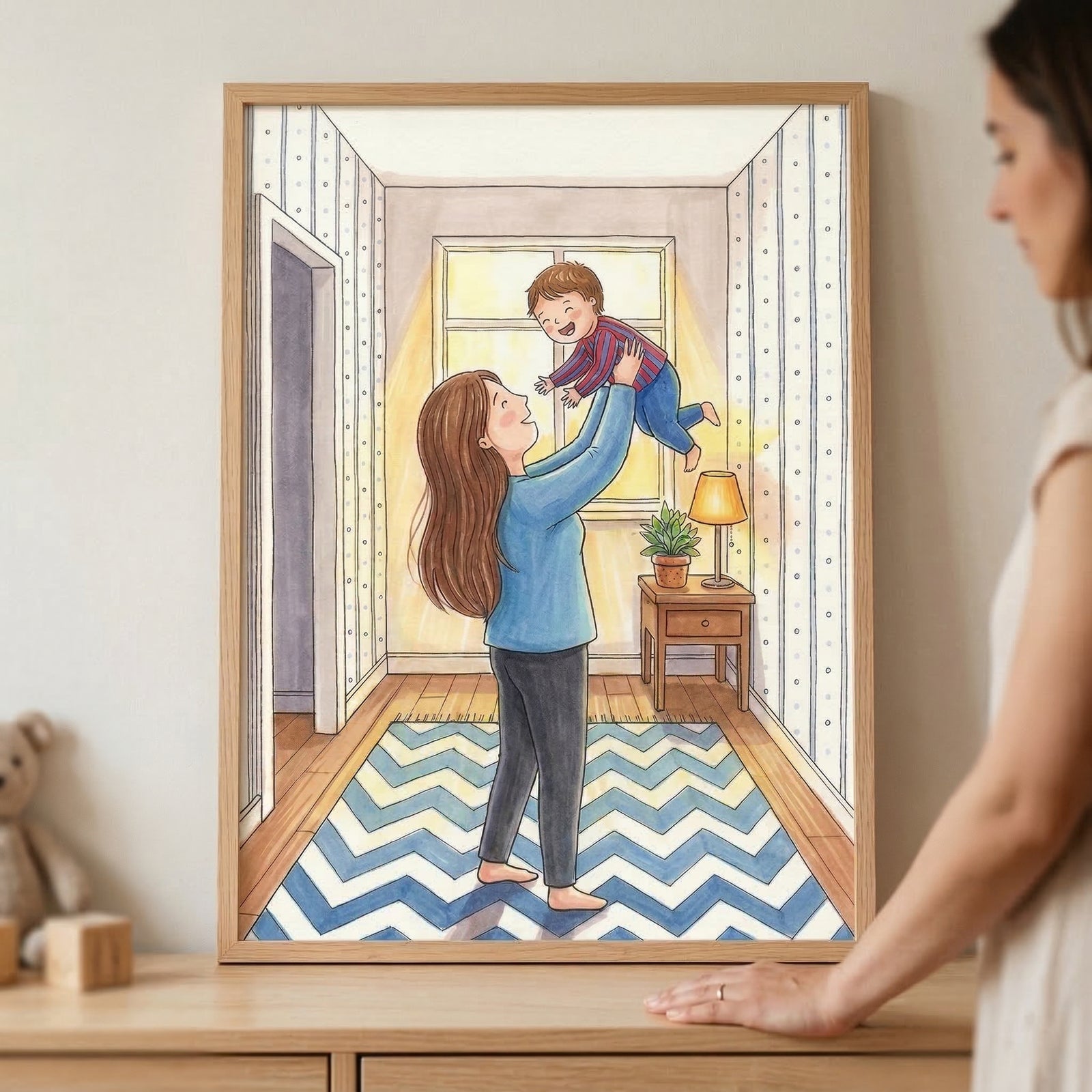 A framed watercolor-style illustration depicts a joyful scene of a woman with long brown hair lifting a small child in a hallway. The woman is wearing a light blue top and dark grey pants, while the child is dressed in a red and blue striped sweater and blue pants. They are standing on a blue and white chevron-patterned rug. 