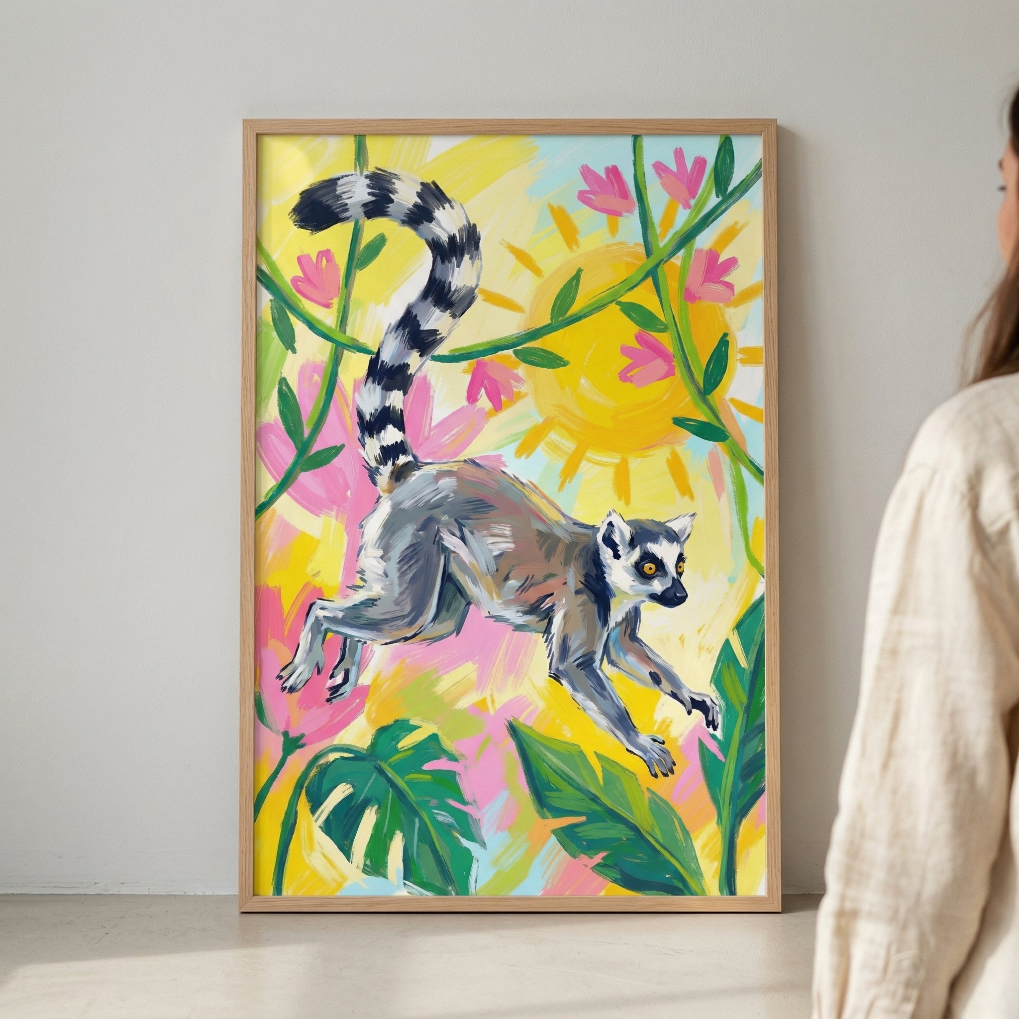 A framed piece of vibrant artwork displays a ring-tailed lemur in a mid-air leap. The lemur, with its characteristic long, black-and-white banded tail, is the central focus against a bright, colorful background. Stylized tropical elements like large green leaves, pink flowers, and a glowing yellow sun surround the animal. The style features bold, expressive brushstrokes and a playful use of color. The art is presented in a simple wooden frame, leaning against a light-colored wall.
