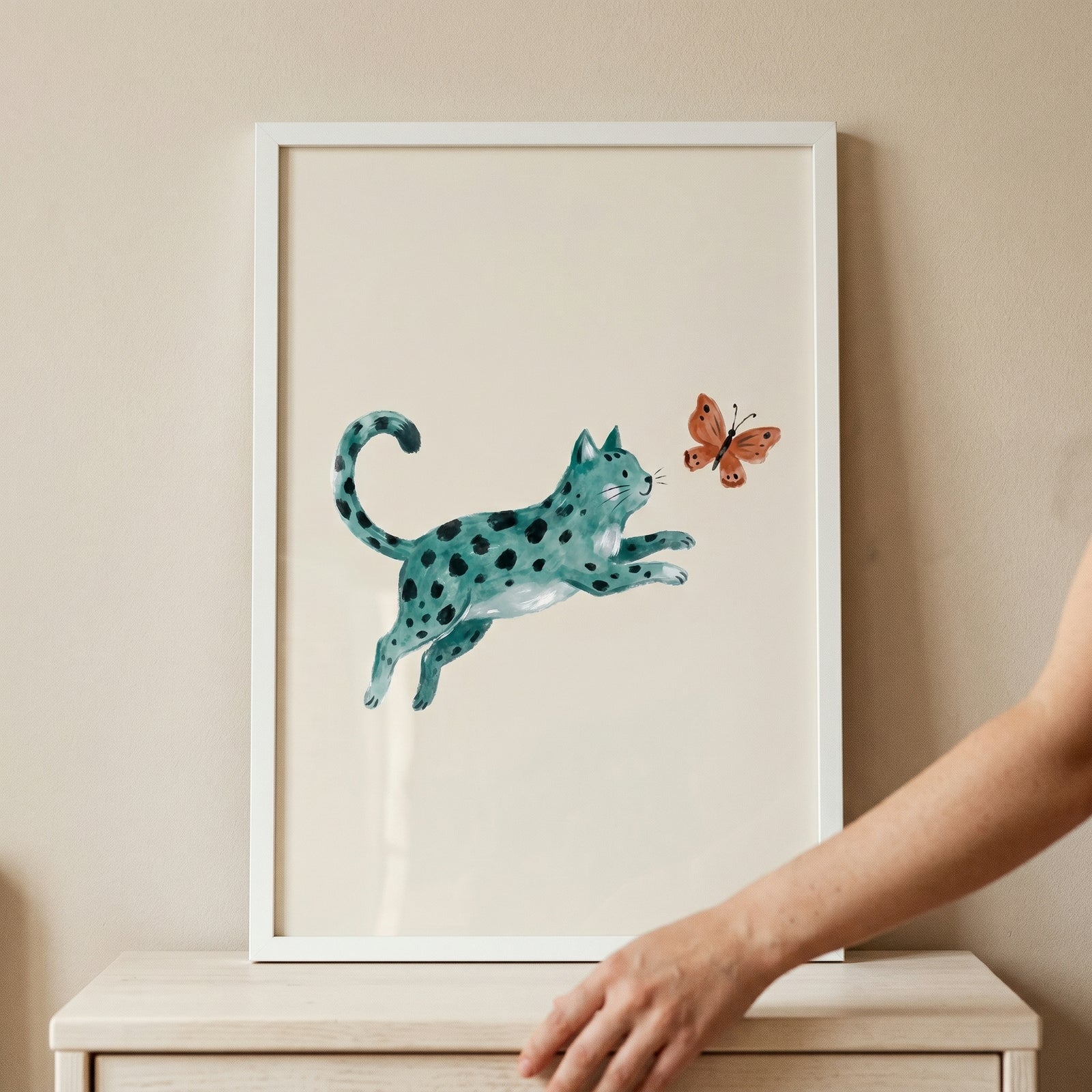 Framed jumping teal spotted cat wall art on a dresser shows the feline pouncing toward an orange butterfly.