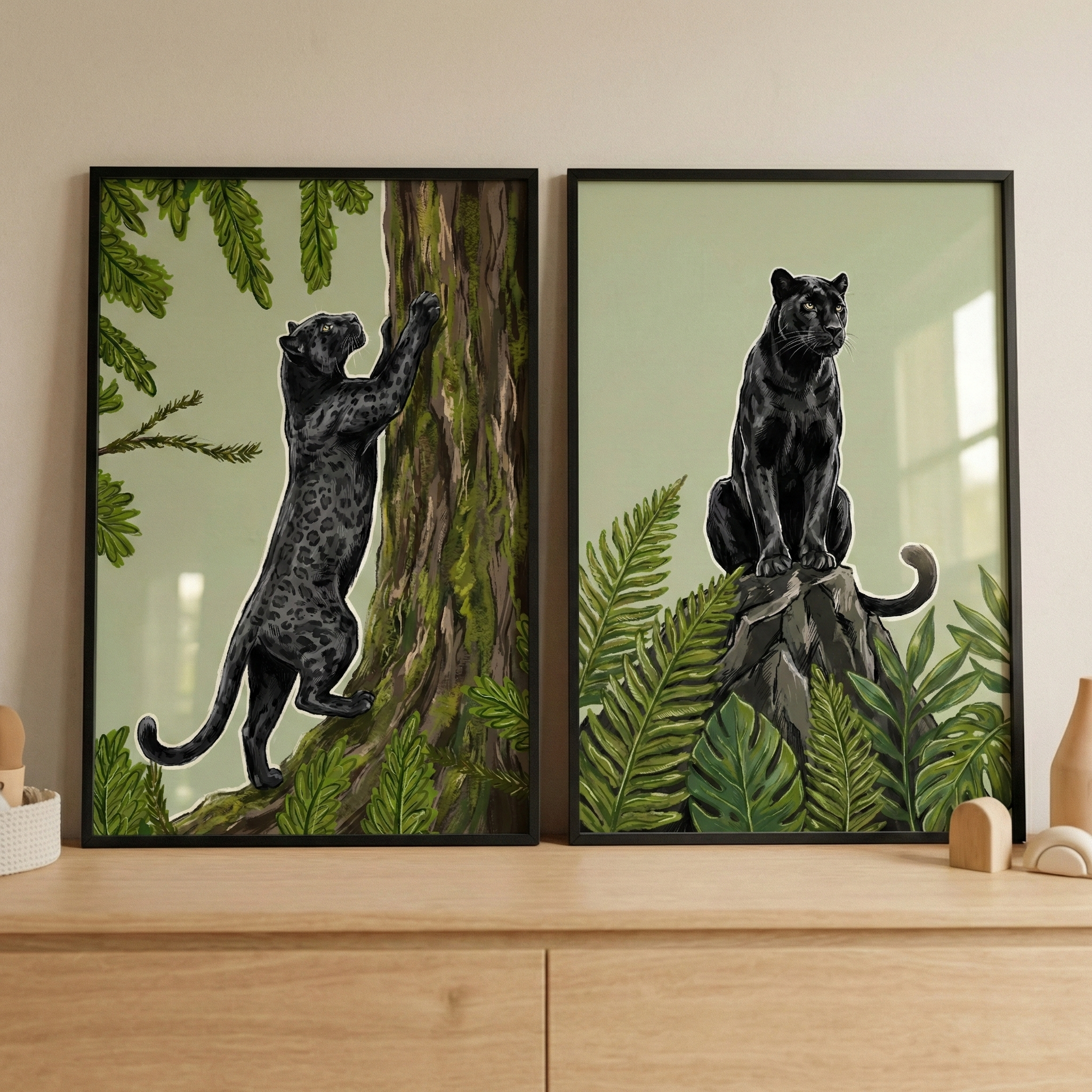 Two thin black-framed illustrations rest side-by-side on a light wood dresser. The left piece shows a black leopard climbing a mossy tree trunk amidst green foliage. The right piece depicts the same animal sitting on a rock, surrounded by lush tropical plants like ferns and monstera leaves. Both artworks feature a muted green background. The setting includes a neutral wall and small decorative wooden items on the dresser's surface.