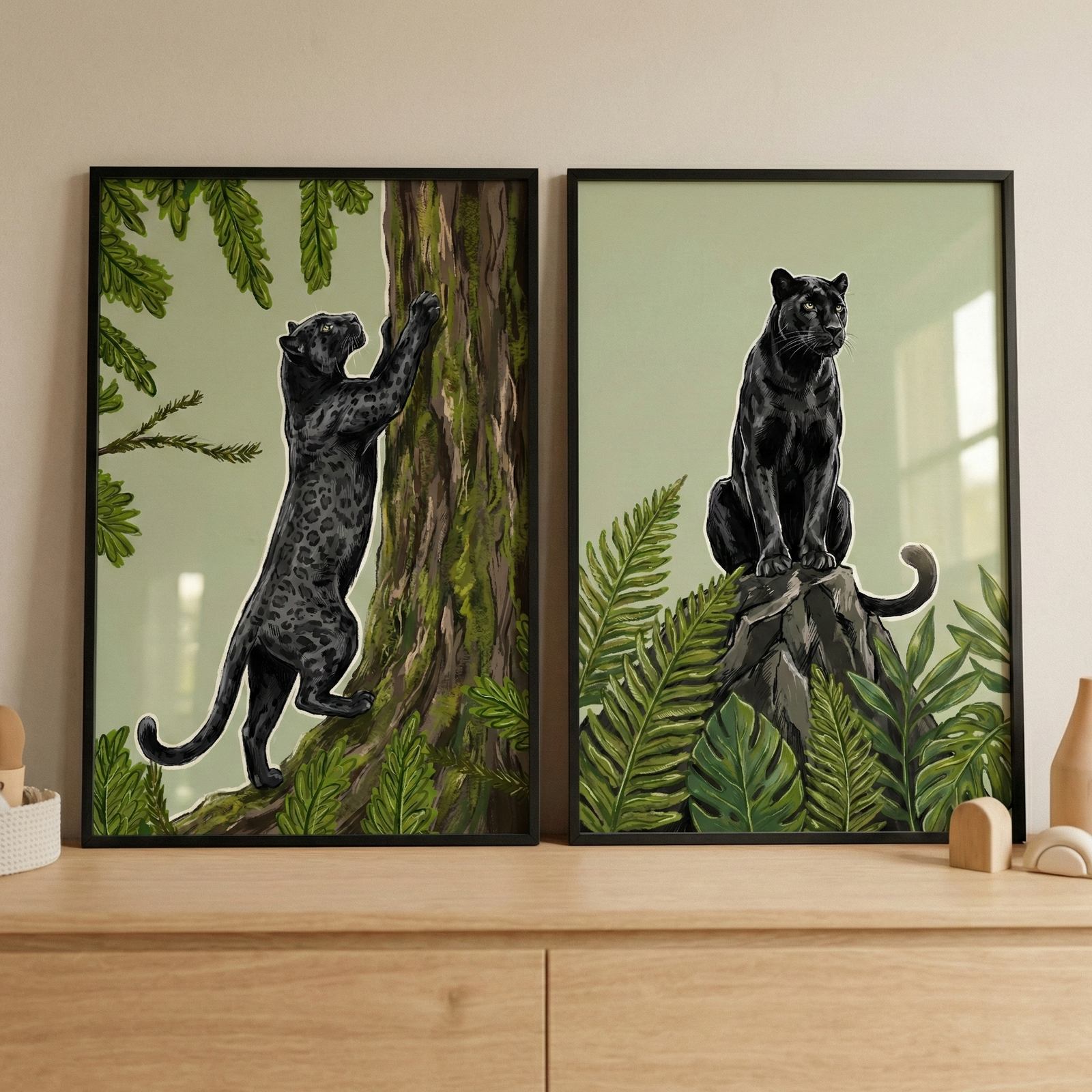 Two thin black-framed illustrations rest side-by-side on a light wood dresser. The left piece shows a black leopard climbing a mossy tree trunk amidst green foliage. The right piece depicts the same animal sitting on a rock, surrounded by lush tropical plants like ferns and monstera leaves. Both artworks feature a muted green background. The setting includes a neutral wall and small decorative wooden items on the dresser's surface.