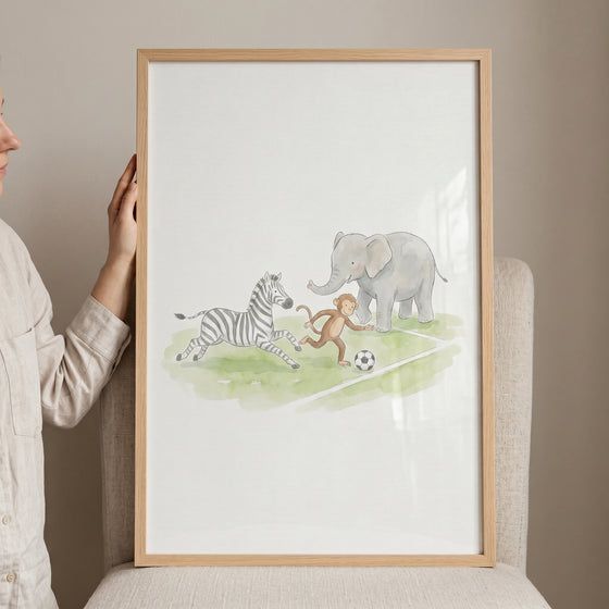 Framed watercolor illustration of a zebra, monkey, and elephant kicking ball brown animals wall art on a field.