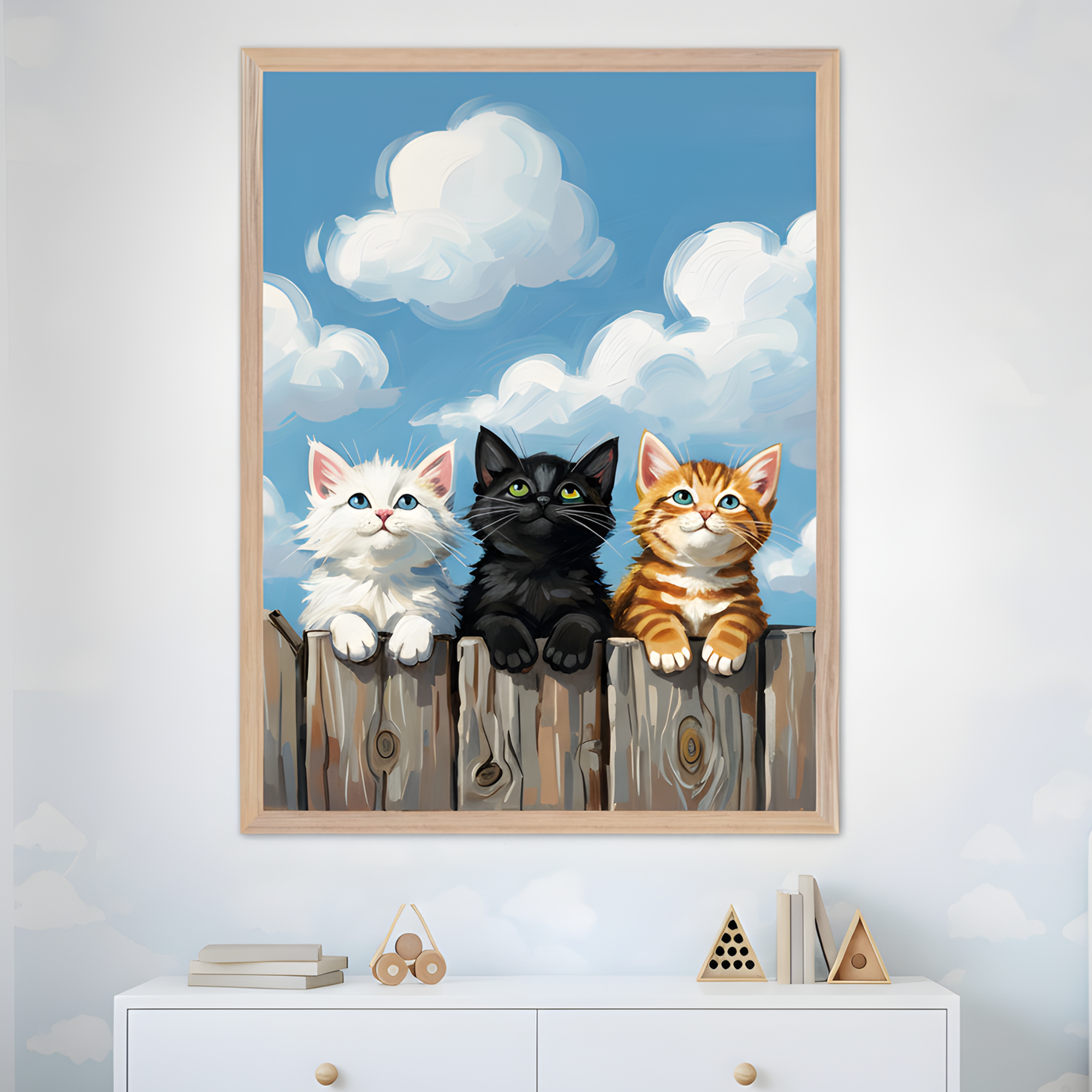 A framed painting of three adorable kittens a fluffy white, a sleek black, and an orange tabby sitting on a wooden fence against a bright blue sky with puffy white clouds. The image adds a playful and cheerful touch to the softly themed nursery, with simple wooden toys and cloud-patterned walls below.