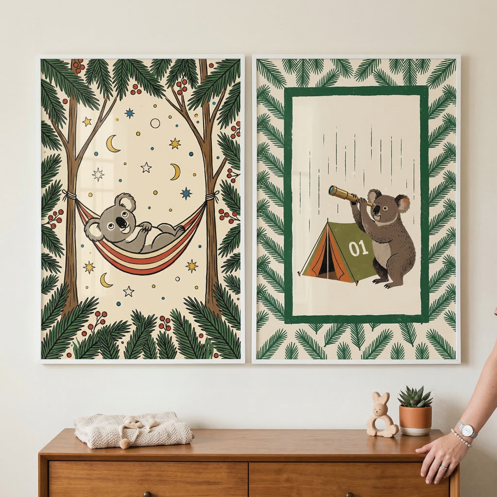 Two framed vertical illustrations hang on a light wall above a wooden dresser. The left piece shows a cartoon koala in a striped hammock between pine trees, surrounded by stars and crescent moons. The right piece features a koala with a telescope next to a green tent under falling rain. Both have pine-themed borders. On the dresser sits a folded cloth, a wooden bunny toy, and a small succulent in a pot. A partial view of a person's arm and hand is on the right side.