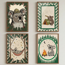 Four framed koala illustrations hang in a two by two grid on a white wall. The top left koala wears a safari hat among pine branches and red berries. The other prints show a koala looking through a telescope beside a tent, relaxing in a hammock under stars and moons, and sitting by a campfire surrounded by pine trees.