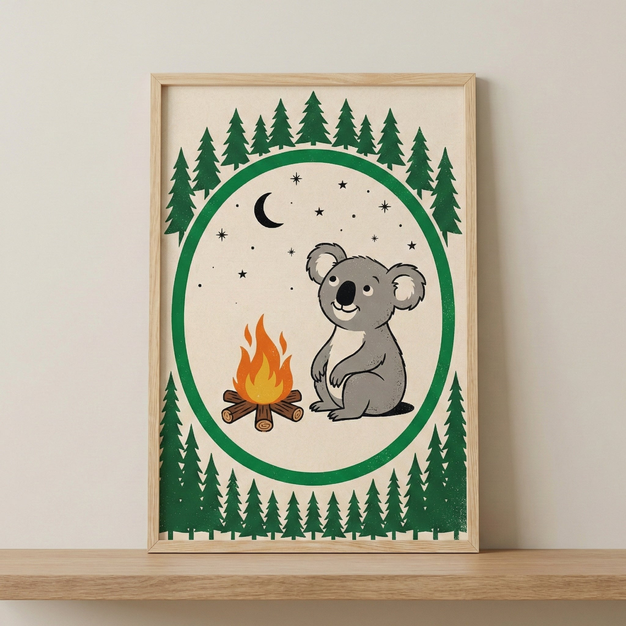 A framed illustration of a smiling cartoon koala sitting next to a small campfire. The central image is set against a cream background and enclosed within a thick green circular border. Above and below the circle are rows of dark green stylized evergreen trees. Inside the circle, a black crescent moon and several small stars are scattered in the sky. The artwork is held in a light wood frame resting on a natural wood surface against a neutral wall.