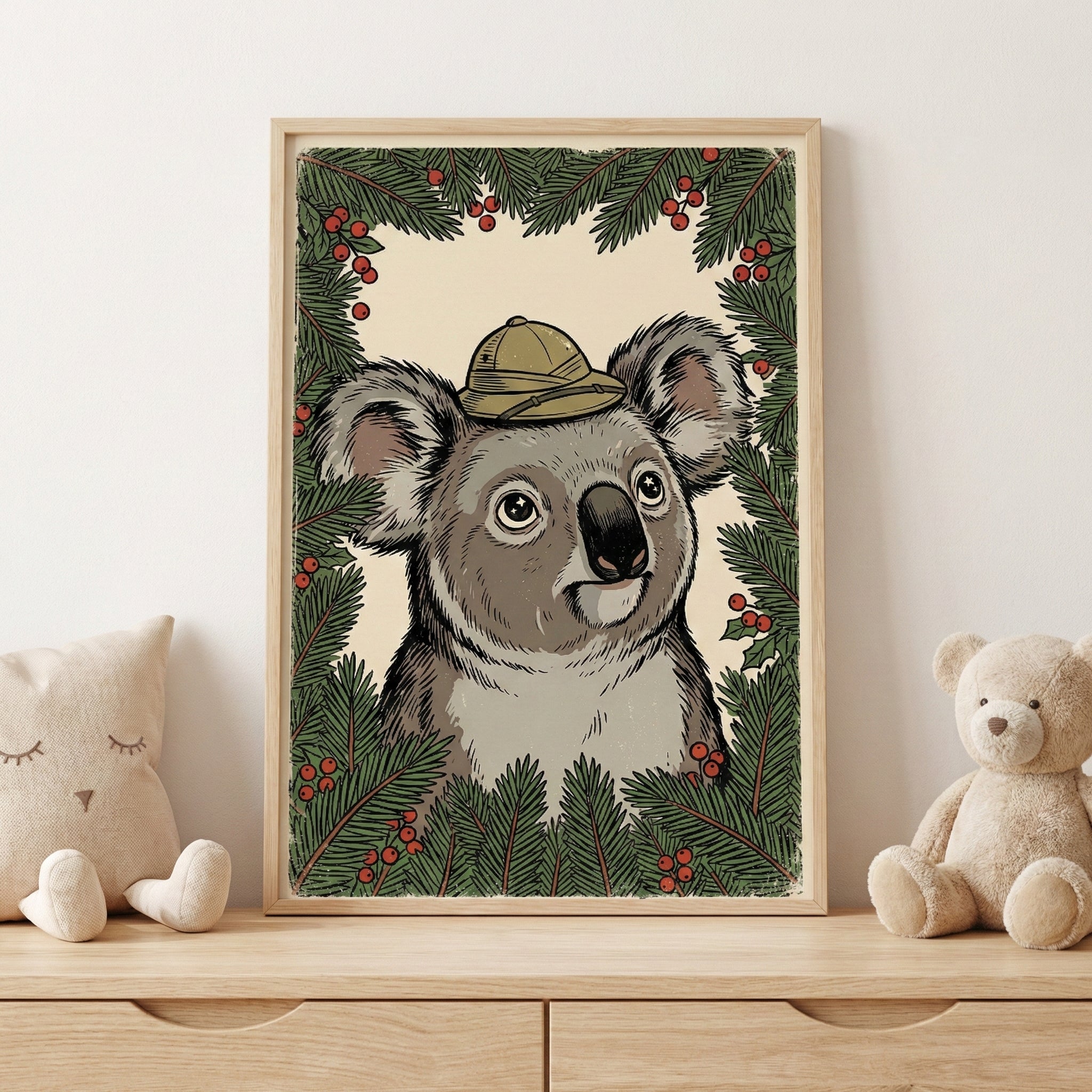 A framed illustration of a koala wearing a tan pith helmet, centered within a border of pine branches and red berries. The artwork has a vintage feel and is displayed on a wooden surface next to a star-shaped plush pillow and a teddy bear.