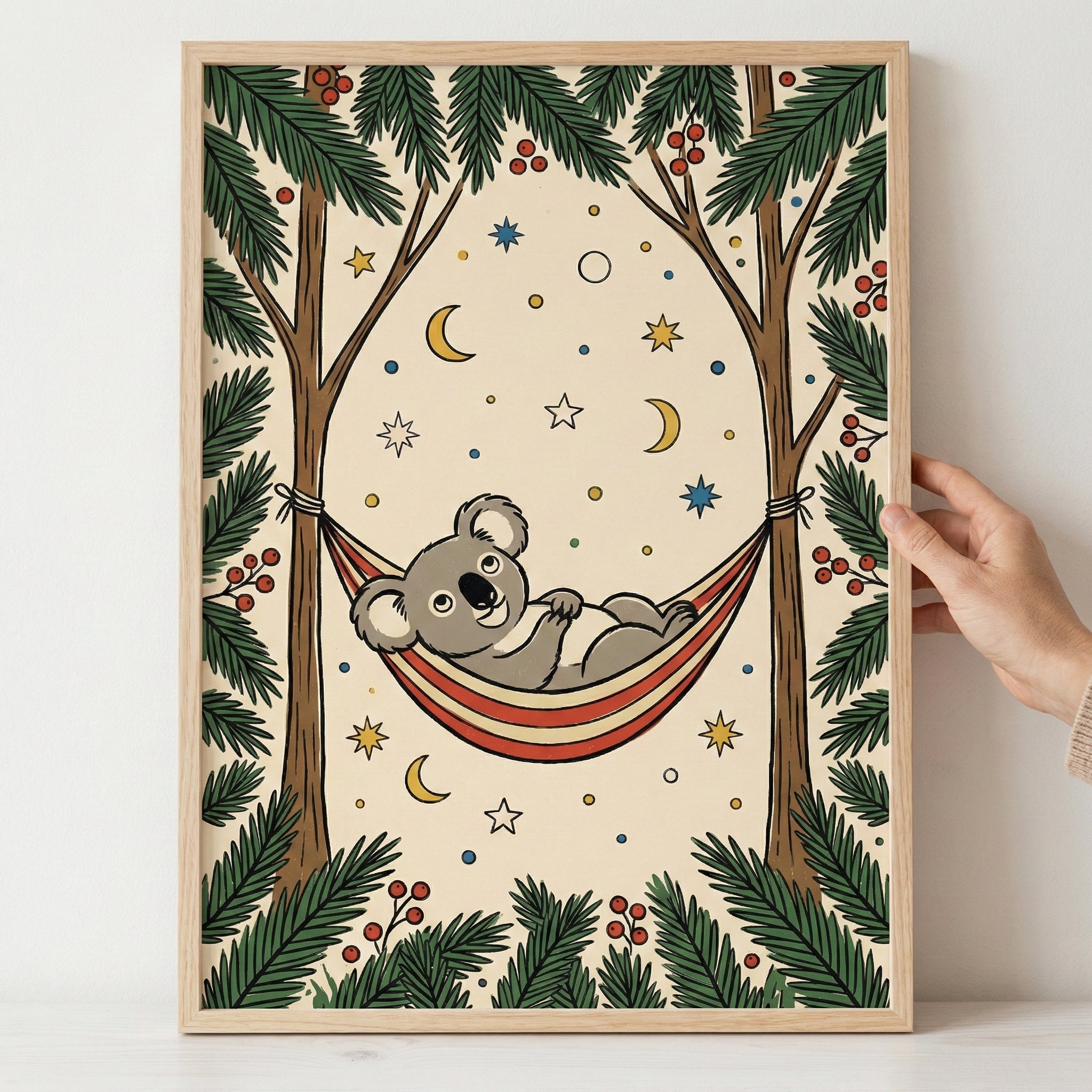 A framed whimsical illustration of a smiling cartoon koala relaxing in a red, yellow, and white striped hammock. The hammock is suspended between two tall trees with green, feathery branches and small red berries. The cream-colored background is decorated with yellow crescent moons, colorful stars, and small dots. A hand holds the right side of the light wood frame against a neutral background.