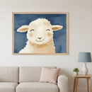 Modern living room with a cream sofa and pink cushion. A large framed, painterly illustration in a light wooden frame of a smiling white lamb with pink cheeks on a dark blue background hangs above the sofa. A side table with a blue lamp and plant is on the right.