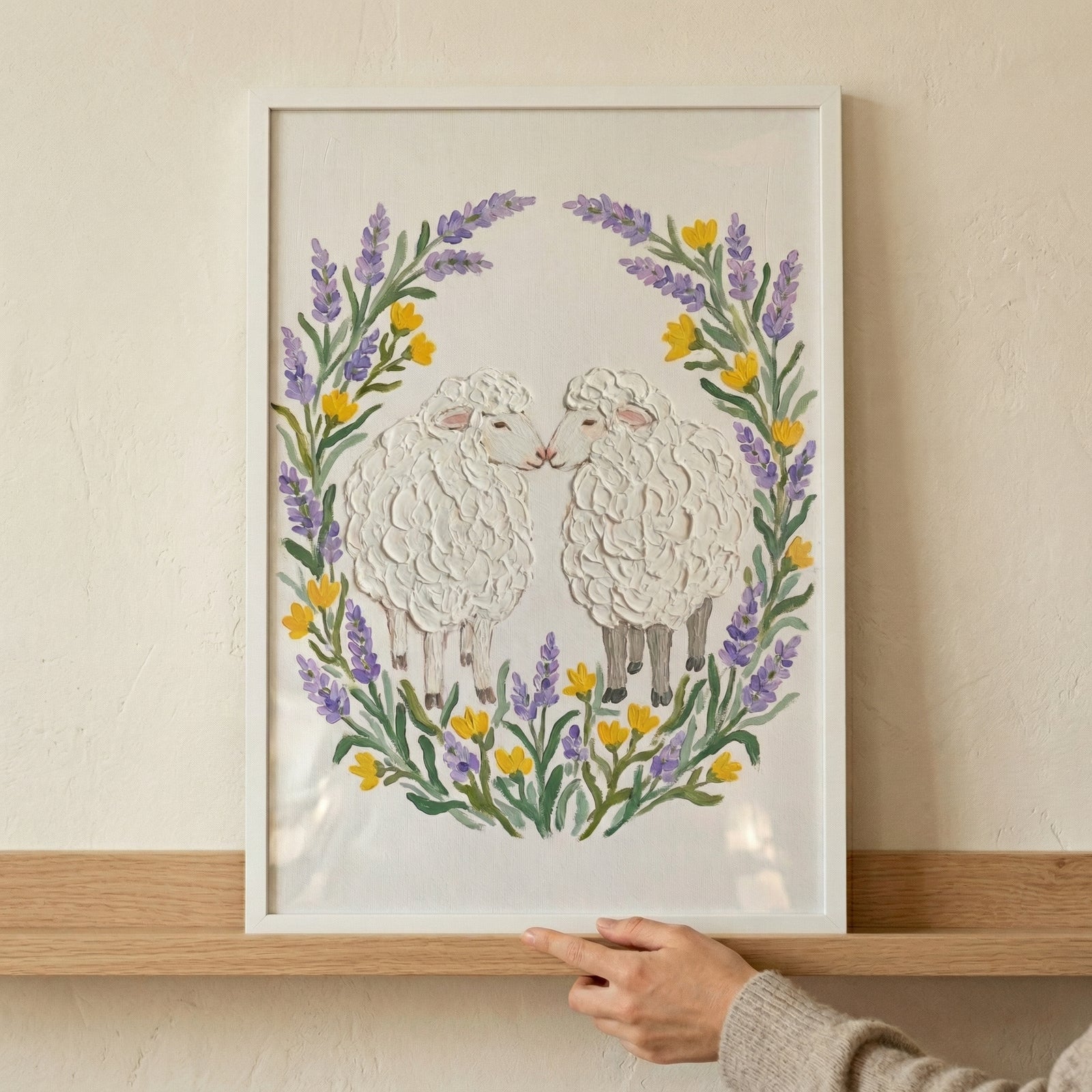 Framed painting of two fluffy sheep facing each other nose to nose surrounded by a circular wreath of purple lavender and small yellow flowers. A hand rests on the wooden shelf beneath the artwork emphasizing the soft textured brushstrokes and cozy decorative style.