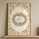 Framed vintage style illustration of two rabbits curled together in a nest of twigs and lavender, surrounded by delicate floral linework and an oval border. The soft neutral tones and detailed design create a peaceful and nurturing focal point for a nursery space.