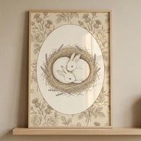 Framed vintage style illustration of two rabbits curled together in a nest of twigs and lavender, surrounded by delicate floral linework and an oval border. The soft neutral tones and detailed design create a peaceful and nurturing focal point for a nursery space.