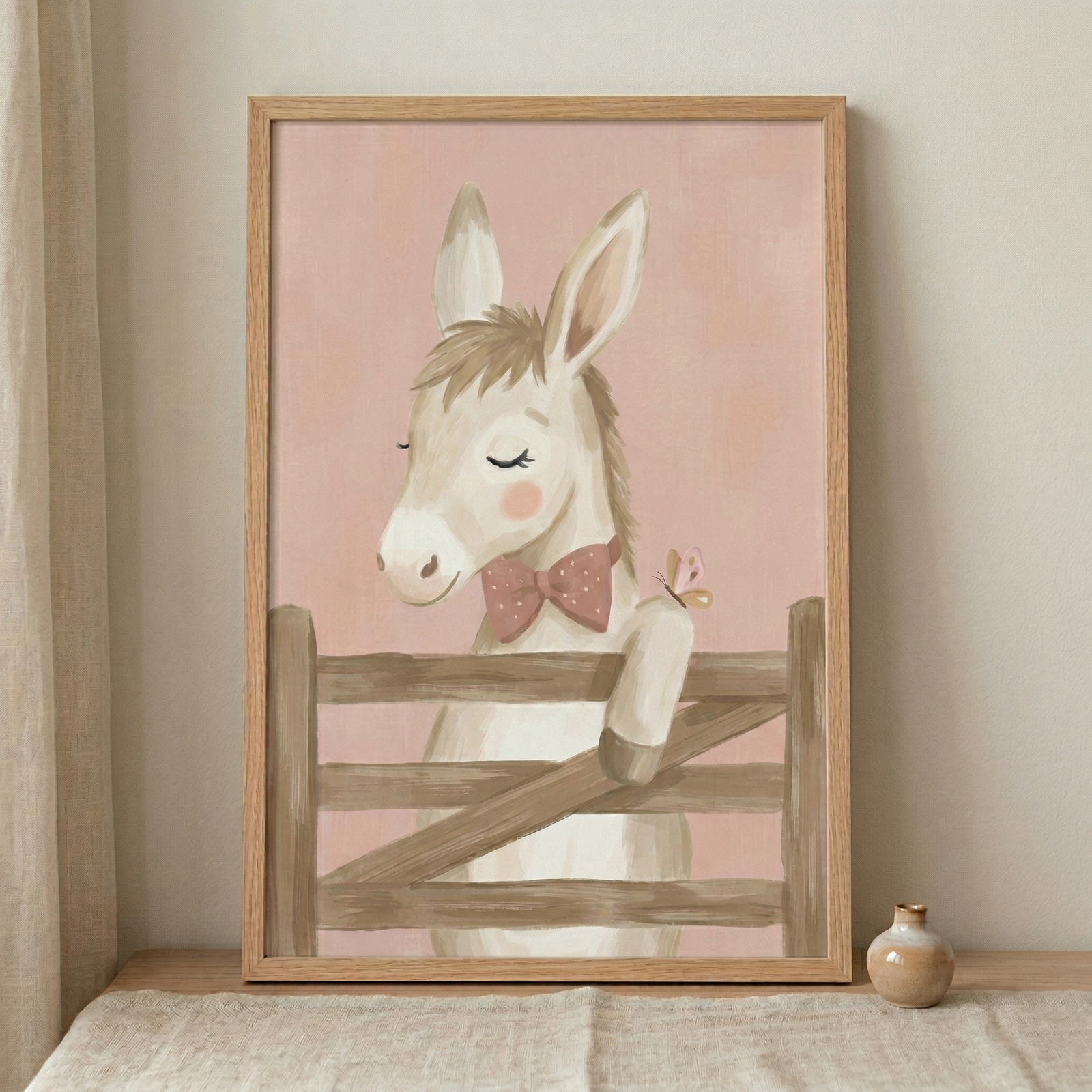 A framed whimsical illustration of a cute young donkey. The donkey, rendered in soft cream and tan tones, has its eyes gently closed and features rosy pink cheeks. It is adorned with a small pink bow tie decorated with white polka dots. The donkey is shown leaning over a simple, rustic wooden fence. A tiny pink butterfly is perched on its front leg. The entire scene is set against a solid, soft pink background with a subtle textured finish. 