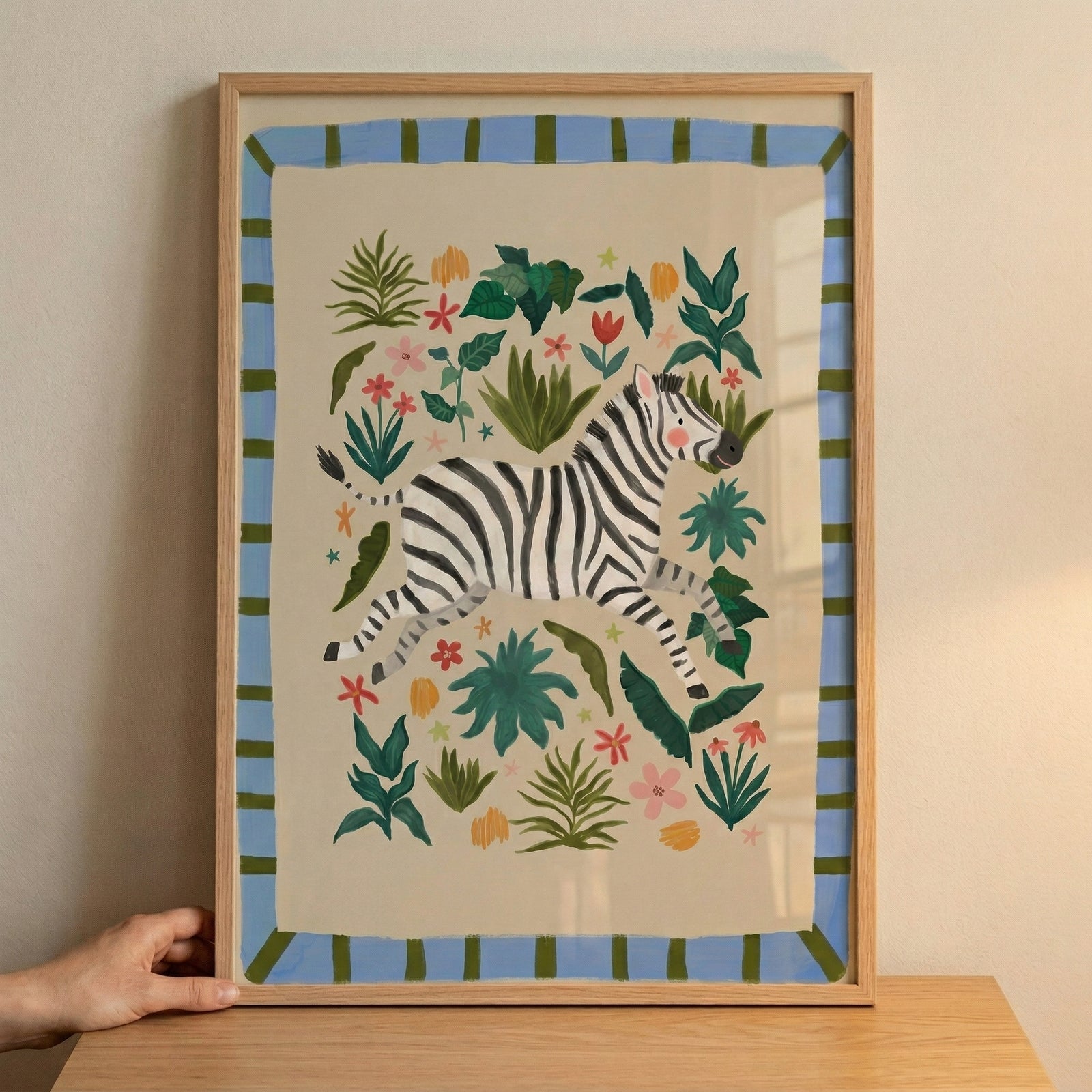 Framed zebra illustration with black and white stripes surrounded by colorful leaves and flowers, styled as playful safari nursery wall art. A hand steadies the frame on a wooden surface, showing scale and placement for decorating a child’s room.