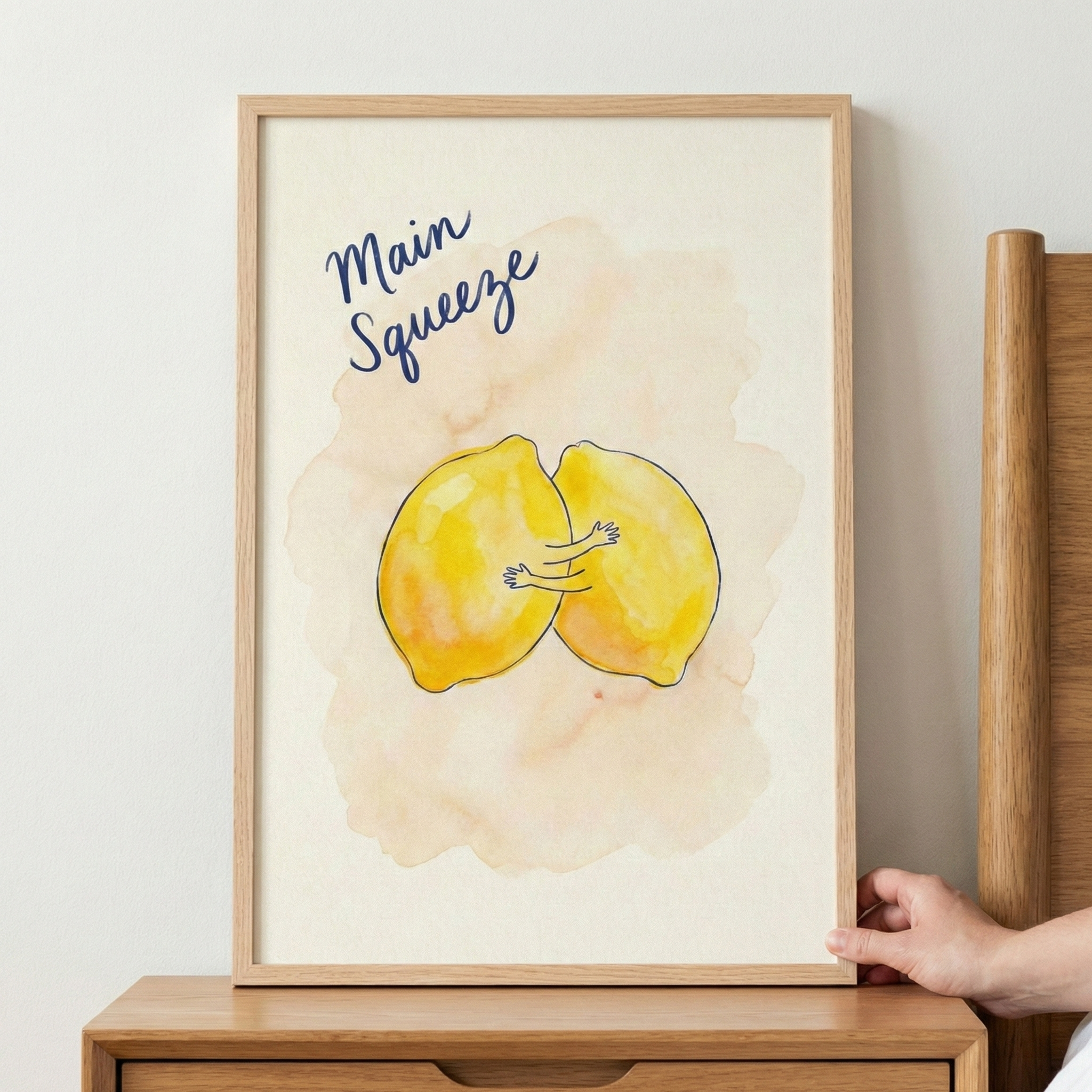 A piece of wall art in a light wooden frame rests on a wooden surface against a plain white wall. The artwork depicts two yellow lemons with simple line-drawn arms hugging each other. Above the lemons, the phrase "Main Squeeze" is written in a dark blue cursive font. The lemons are set against a background of soft peach watercolor-style washes. A hand is seen touching the right edge of the frame.