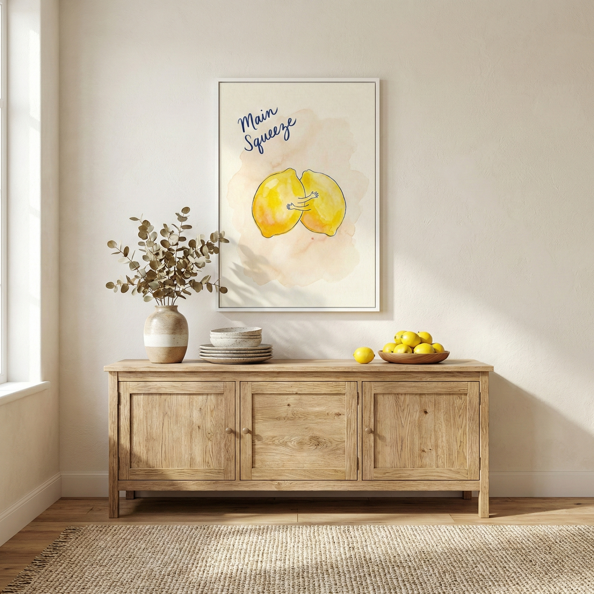 A brightly lit, minimalist interior featuring a light-toned wooden sideboard against a plain, off-white wall. On the sideboard sits a textured ceramic vase filled with dried branches, a stack of neutral-colored ceramic plates and bowls, and a wooden bowl containing several lemons. A single lemon rests on the surface next to the bowl. 