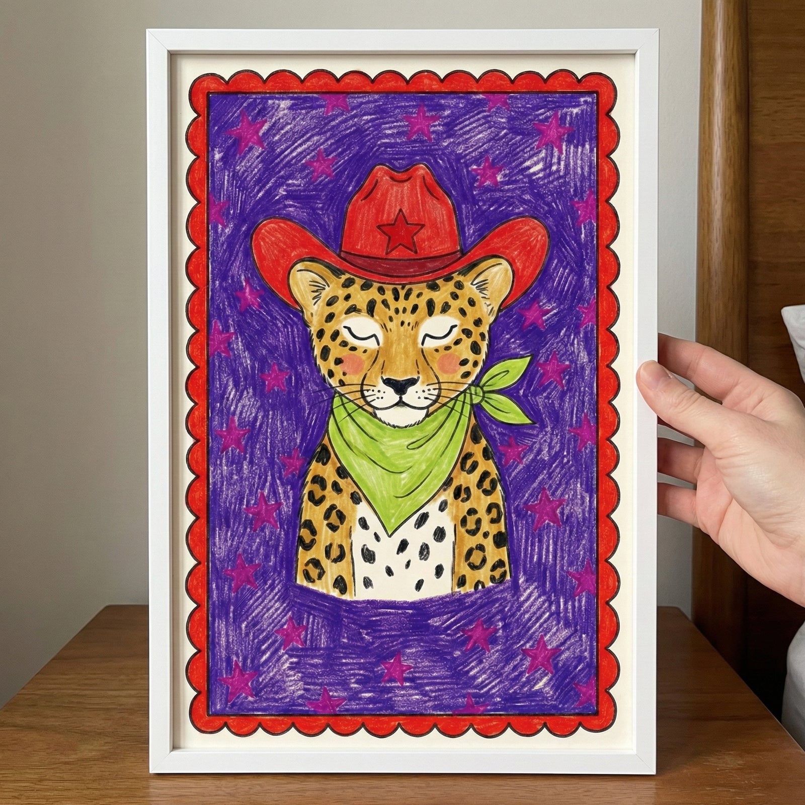 Colorful framed illustration of a leopard wearing a red cowboy hat with a star and a green bandana, set against a purple background with pink stars and a red scalloped border. A hand holds the framed art above a wooden surface.