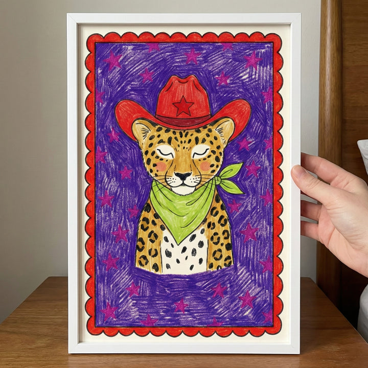 Colorful framed illustration of a leopard wearing a red cowboy hat with a star and a green bandana, set against a purple background with pink stars and a red scalloped border. A hand holds the framed art above a wooden surface.