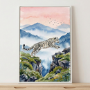 A framed watercolor illustration of a snow leopard jumping between two rocky mountain ledges. The snow leopard has a thick, spotted coat and is captured mid-leap with its long tail extended. The rocky edges are dotted with small white and pink flowers and green mossy foliage. The background features layered, misty blue mountains beneath a soft pink and orange sky where a small flock of birds flies in the distance. The artwork is shown inside a white frame, leaning against a neutral-colored wall on a light w