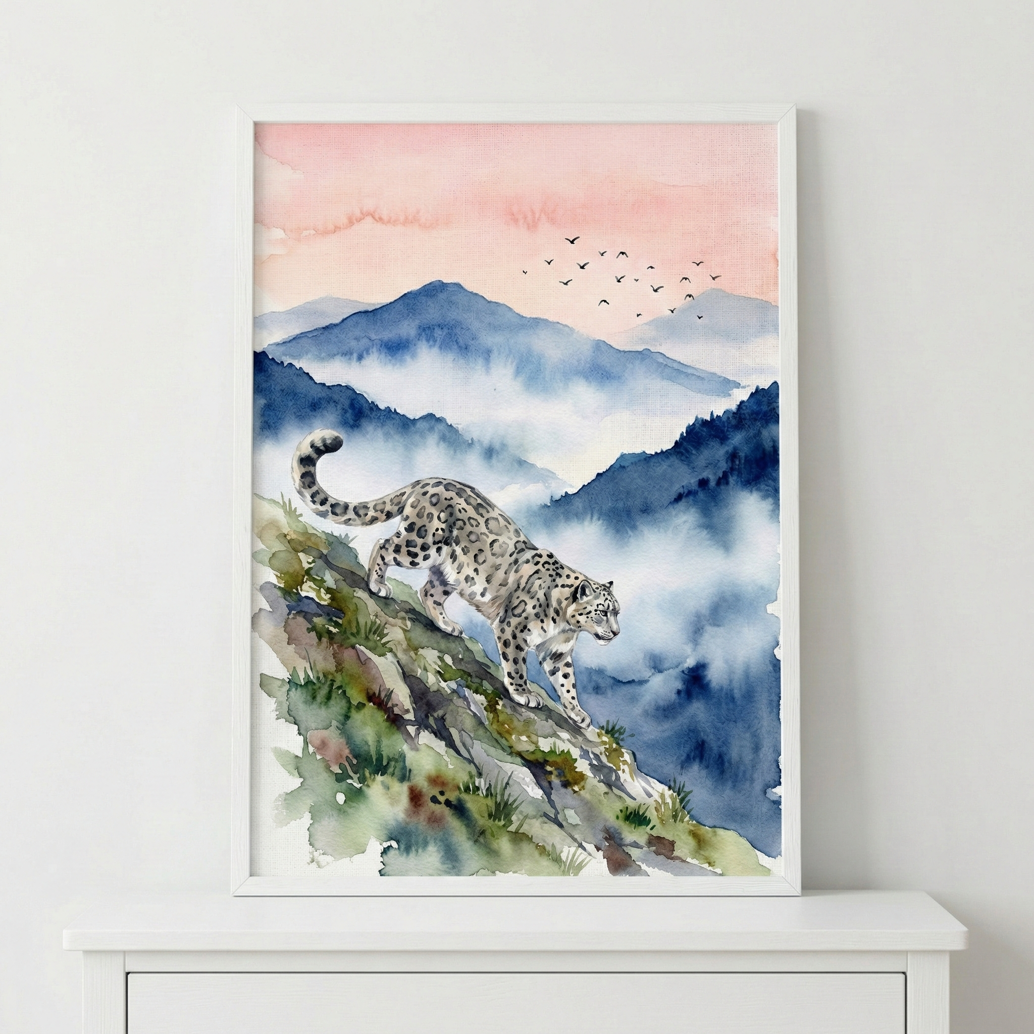 A watercolor illustration shows a snow leopard carefully walking down a rocky, mossy slope. The background features layers of misty blue mountains under a soft pink sky where a flock of birds is flying in the distance. This artwork is displayed in a white frame, resting on a white wooden surface. The style uses soft washes of color to create an atmospheric mountain scene, highlighting the animal's detailed spotted fur and powerful form.