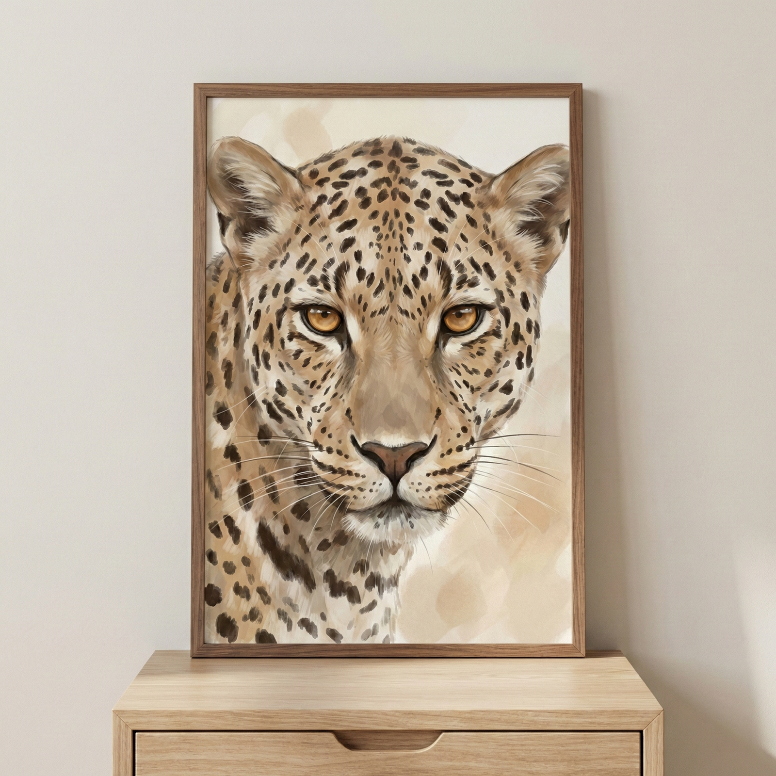 A vertical artwork featuring a detailed, close-up portrait of a leopard's face. The large cat has intense amber eyes and a patterned coat with black rosettes and spots on a light tan background. Delicate, long whiskers fan out from its muzzle. The piece is housed in a simple wooden frame and sits on a light-colored wooden cabinet against a plain, off-white wall. The style is illustrative and realistic with soft, textured details.
