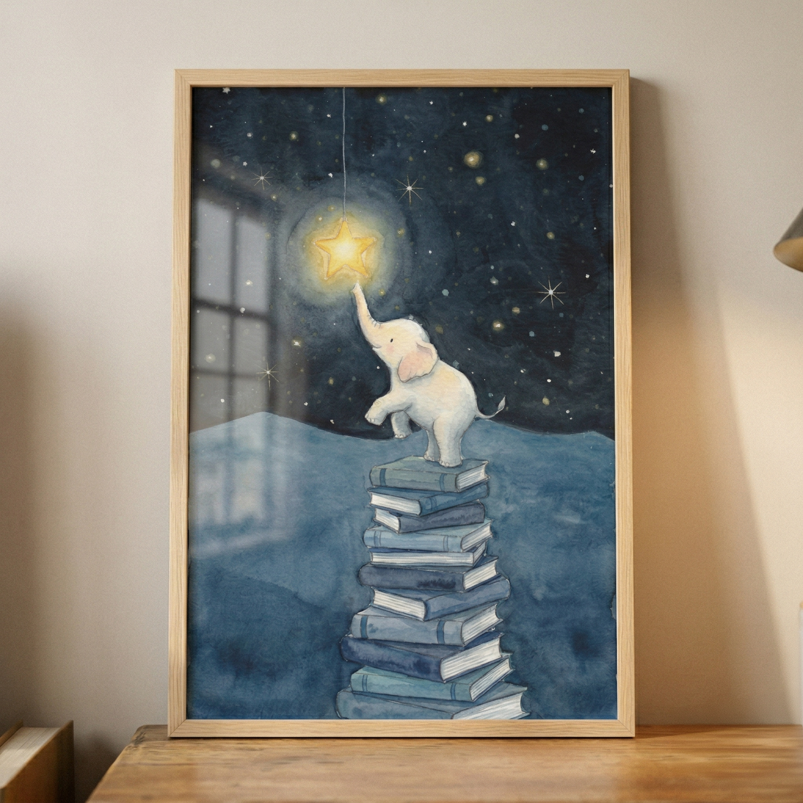 This framed illustration depicts a small, white cartoon elephant perched atop a tall stack of blue books. The elephant reaches its trunk upward to touch a single, large, glowing yellow star suspended from a thin line. The scene is set against a deep blue night sky filled with twinkling stars and a soft, silhouetted mountainous landscape in the distance. The artwork features a gentle watercolor texture and is presented in a light wood frame resting on a wooden surface.
