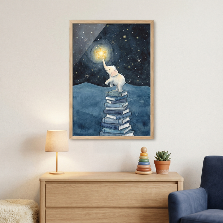 A framed watercolor-style illustration hangs on a light-colored wall above a wooden dresser. The artwork depicts a small, light-pink cartoon elephant standing on its hind legs on top of a tall stack of blue books. The elephant reaches its trunk up toward a large, glowing yellow star in a dark blue night sky filled with smaller stars. Below the frame on the dresser are a small lamp with a beige shade, a wooden ring stacker toy, and a small potted succulent. A blue armchair is partially visible.