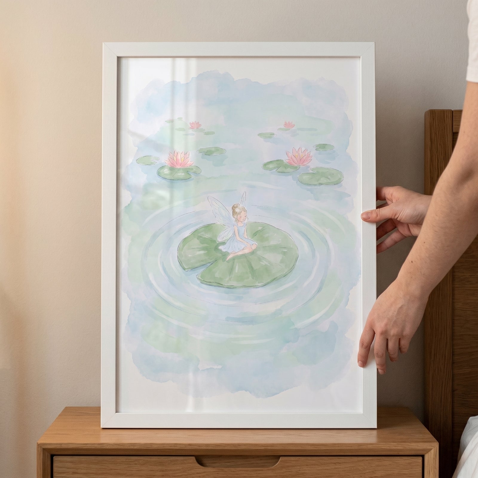 Hands hold a white-framed lily pad fairy wall art of a winged figure on a leaf in a pond with pink water lilies.
