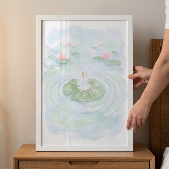 Hands hold a white-framed lily pad fairy wall art of a winged figure on a leaf in a pond with pink water lilies.
