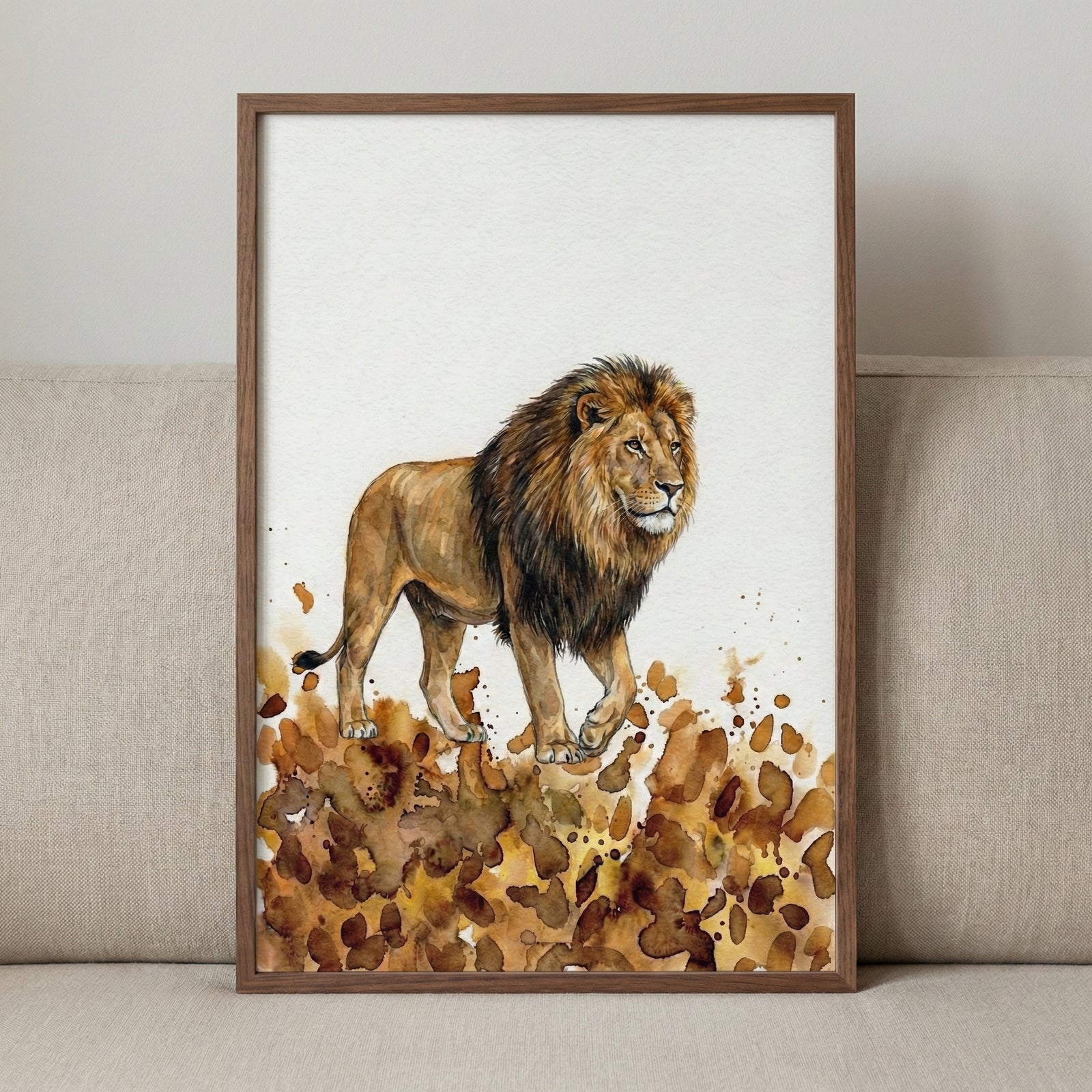 Close-up of a watercolor illustration featuring a lion with a full mane, standing on a bed of golden and brown splashes that evoke natural terrain. The detailed animal artwork is framed in wood and propped on a neutral fabric surface.